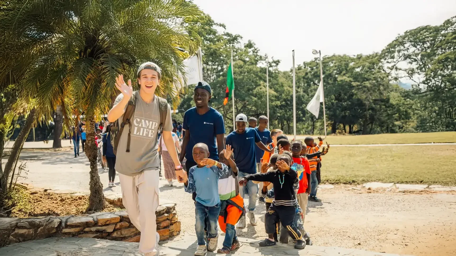 A group of children and young adults walking outdoors, waving, and smiling in a park with trees and flagpoles in the background.