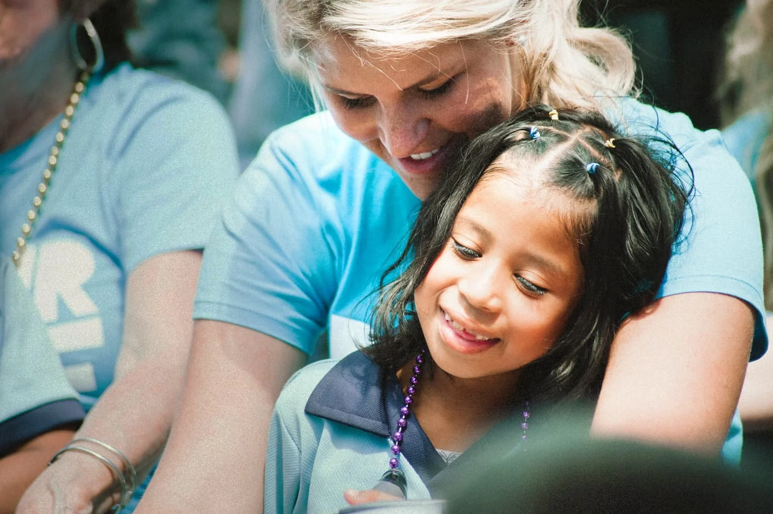 A woman with blonde hair hugging a young girl with dark hair and braids, both smiling