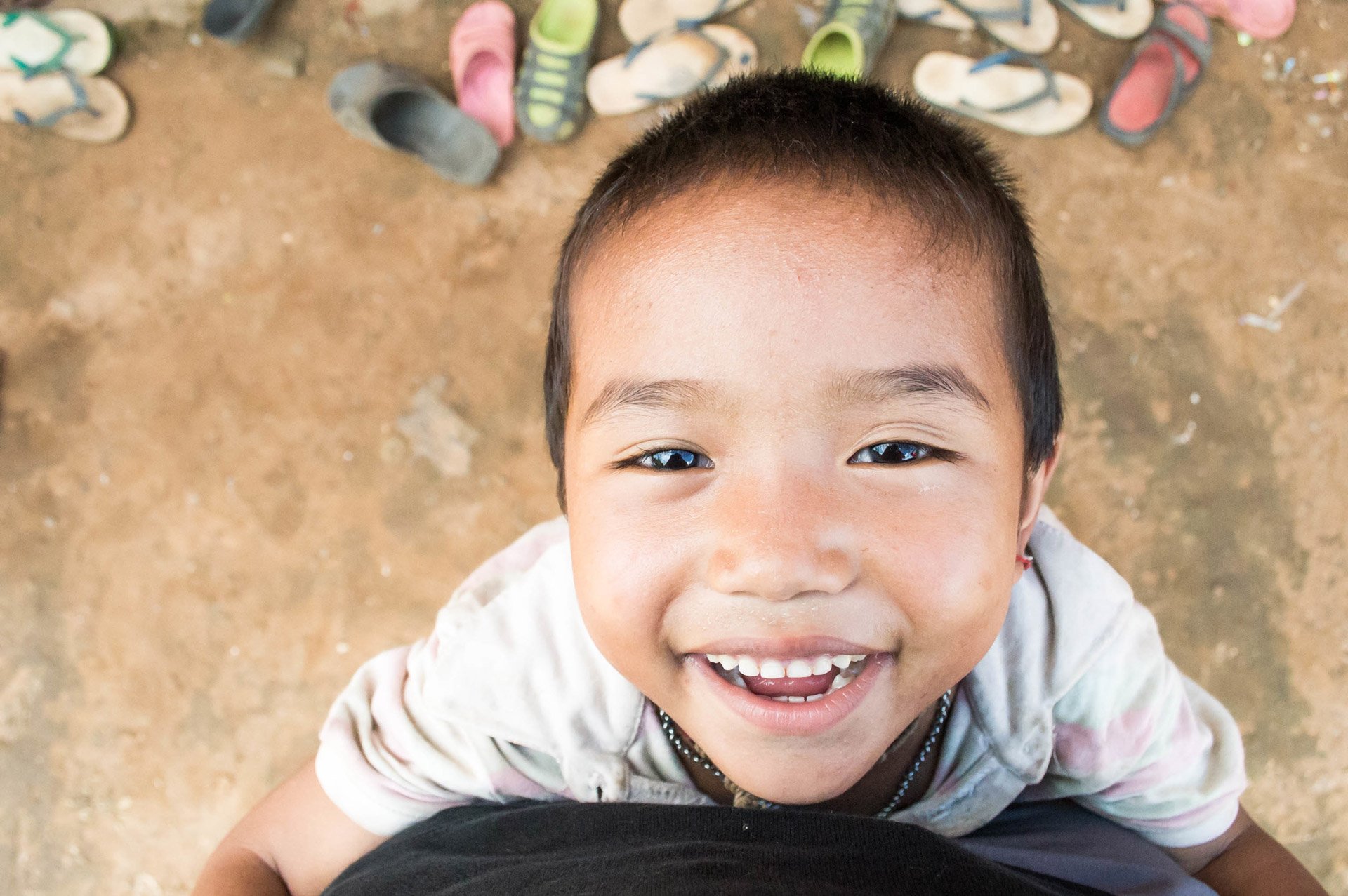 A young boy with short dark hair smiling and looking up at the camera, standing on a dirt ground. Several pairs of sneakers in various colors are visible in the background.