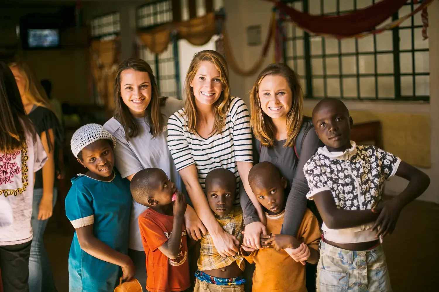 A group of three smiling women and five children of diverse ethnicities posing together indoors.