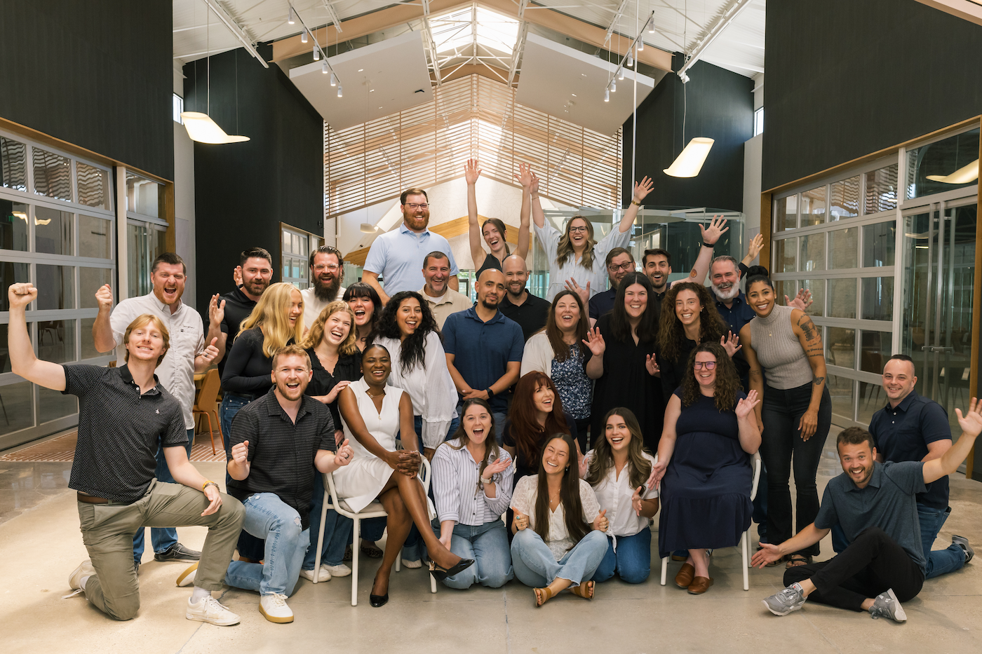 A diverse group of people celebrating and smiling in a modern, well-lit indoor space with large windows and wooden accents.