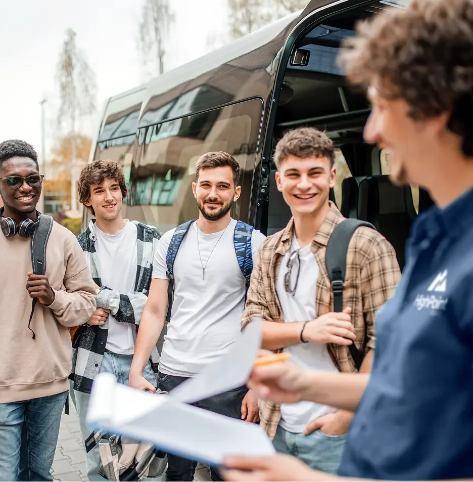 A group of five young men standing outside by a large bus, engaging in conversation, with one man holding a notebook, while a woman with a blue shirt talks to them.