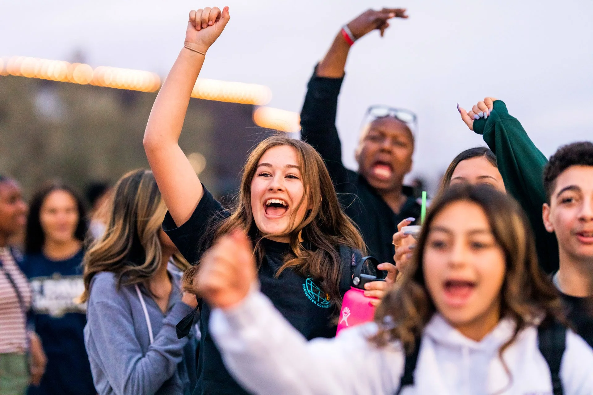 A group of people celebrating outdoors, with one young woman smiling and raising her hand, surrounded by others including a man in the background extending his arm.