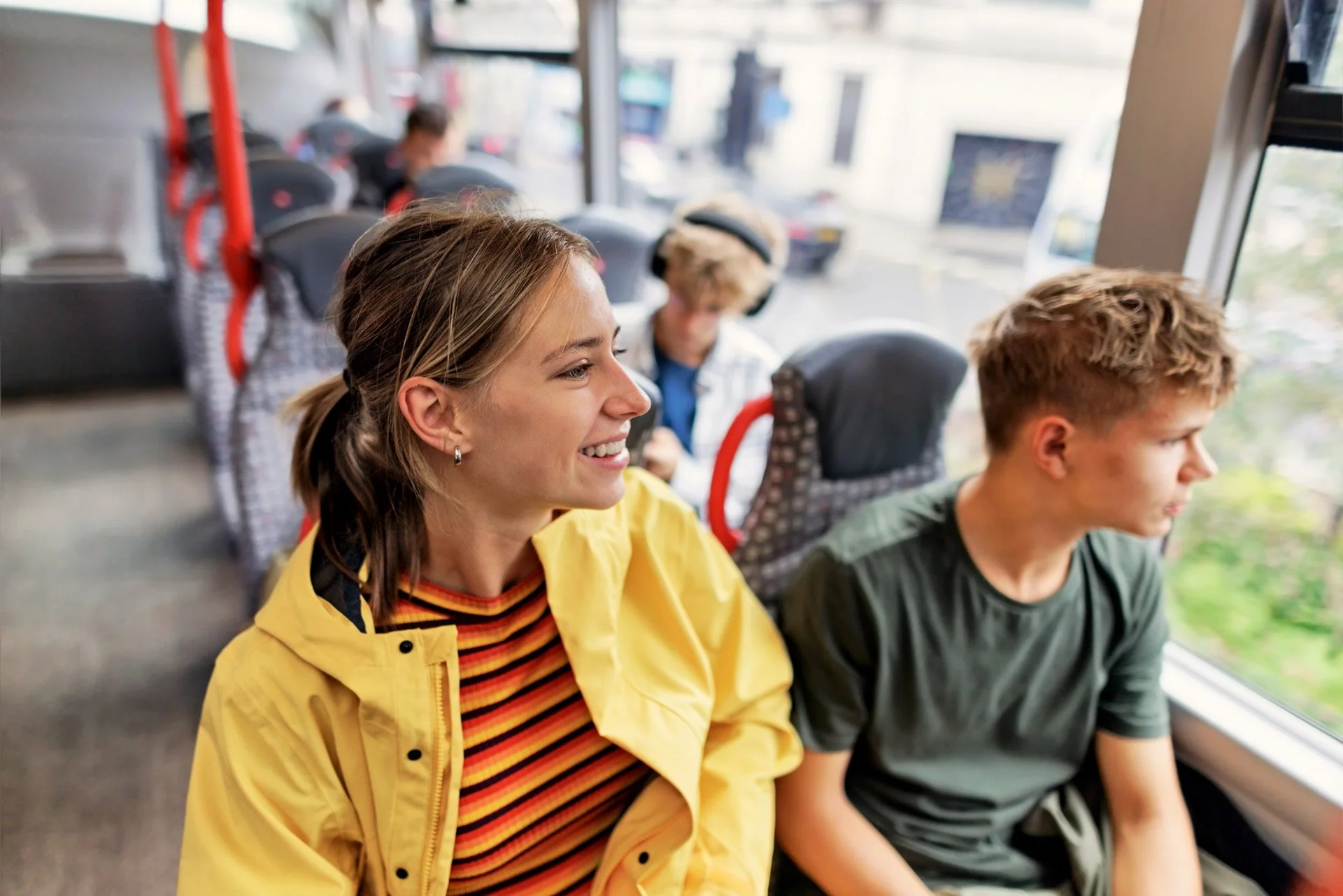 A group of young people sitting on a bus, looking out the window, with a smiling girl in a yellow jacket in the foreground.