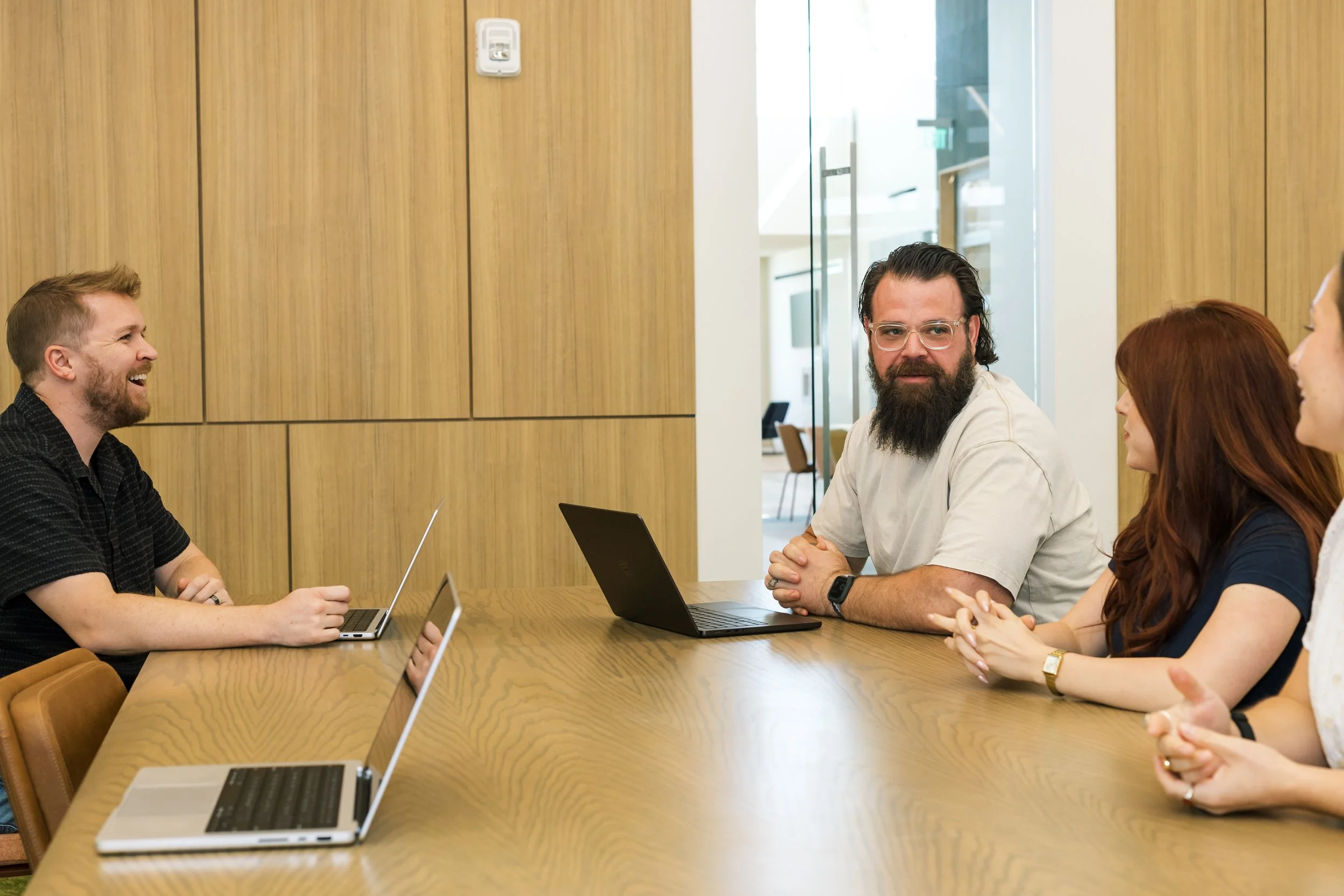 A group of five people sitting around a conference table with laptops, engaged in a discussion in a modern office setting.