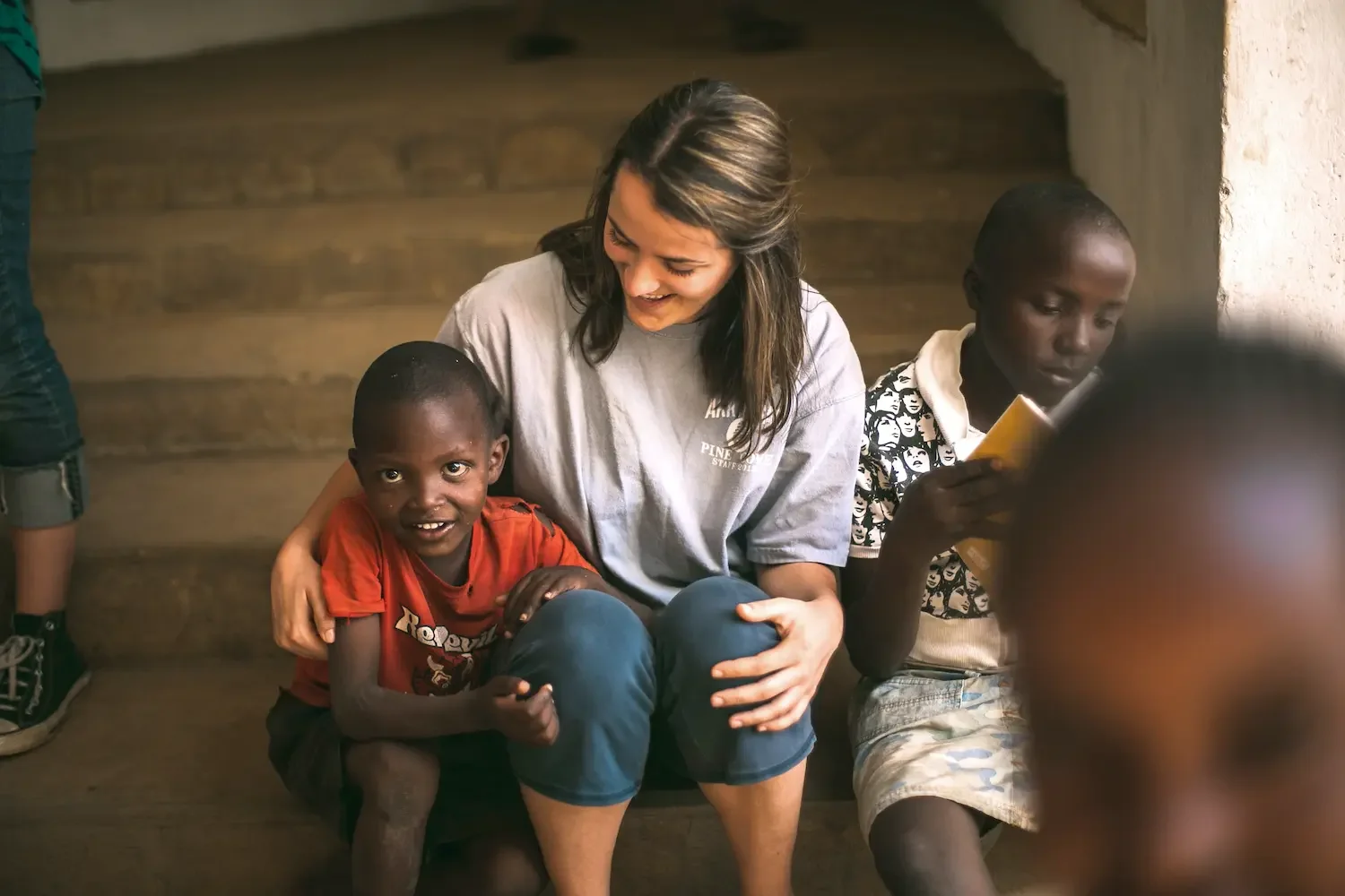 A woman sitting with and hugging a young boy in a red shirt, both smiling at the camera, while another boy with short hair and a patterned shirt reads a book nearby inside a rustic wooden building.