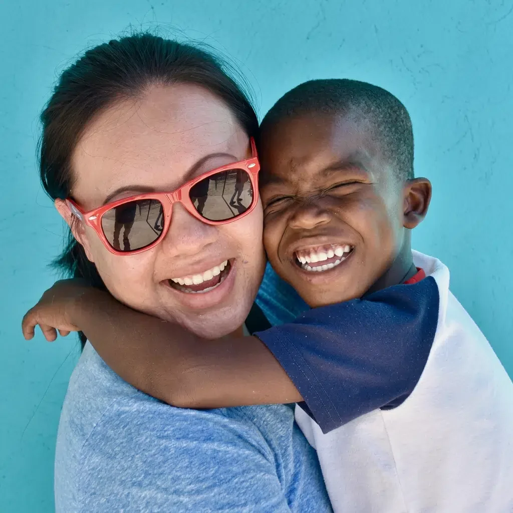 Woman with sunglasses hugging a laughing boy in front of a blue wall.