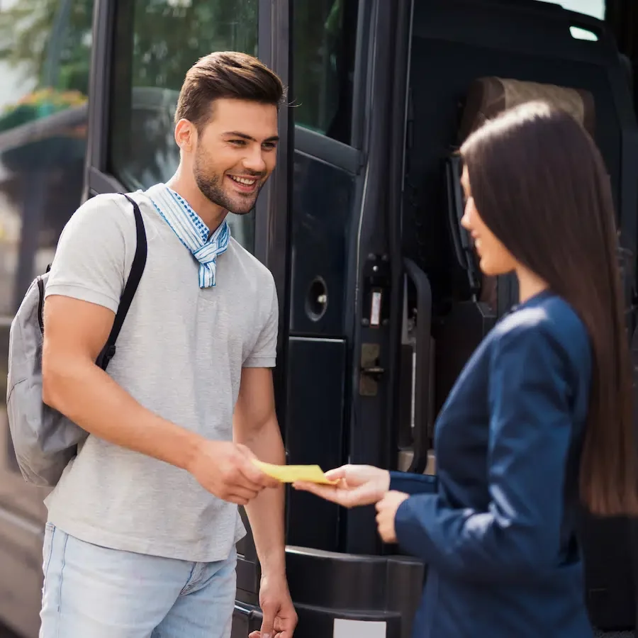 A young man with a backpack receiving a yellow card or paper from a woman in a dark blazer, standing near a bus.