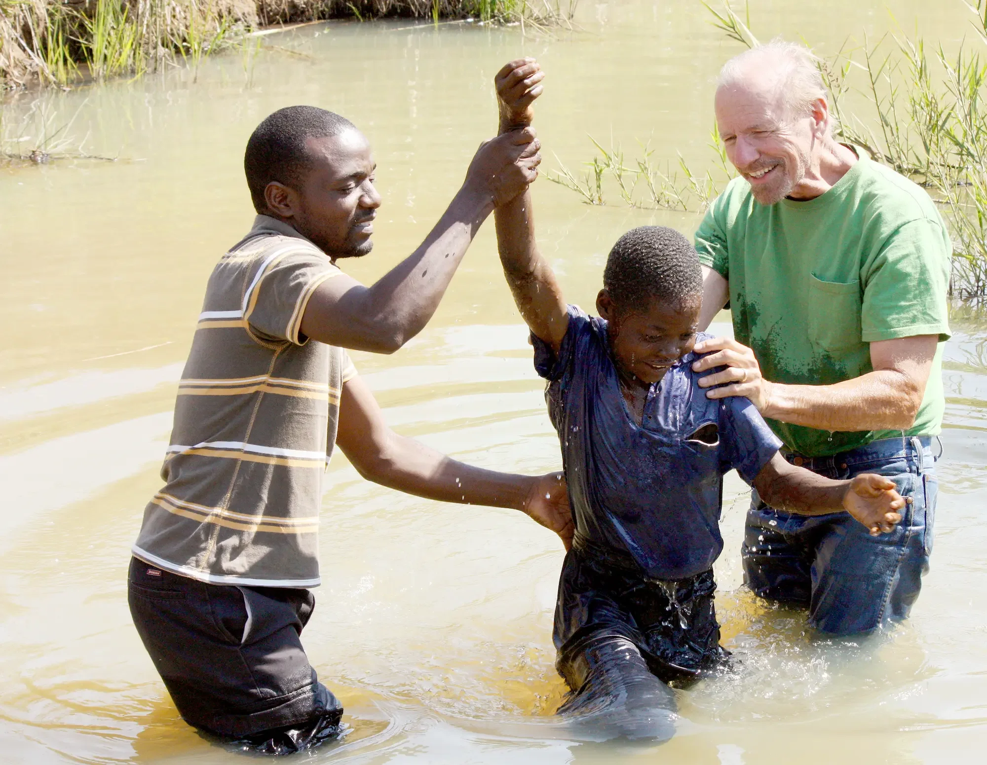 A group of people are in a river participating in a baptism ceremony, with some holding hands and smiling.