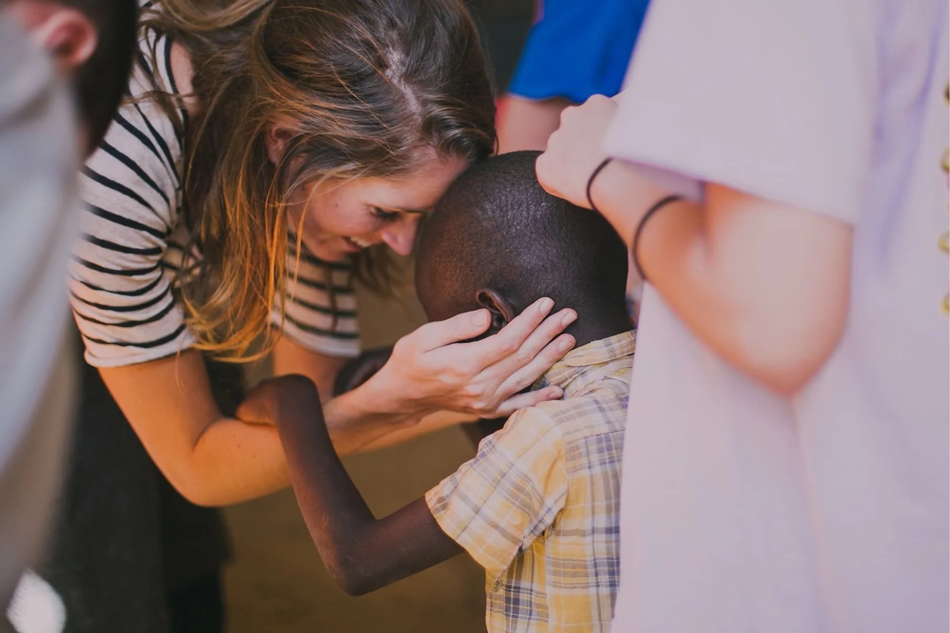 A woman with long, brown hair smiling and leaning close to a young boy, who is seated and has short black hair. The woman is holding the boy's head gently while they both lean in, with several other people standing around them, partially visible.