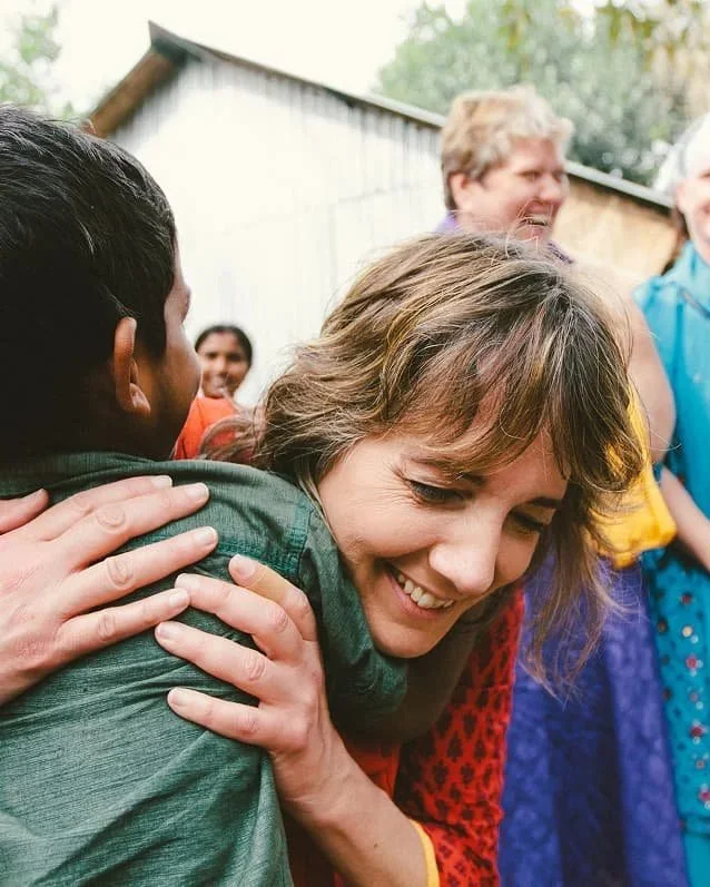 A woman is hugging a young boy outdoors, surrounded by several other smiling people, with trees and a building in the background.