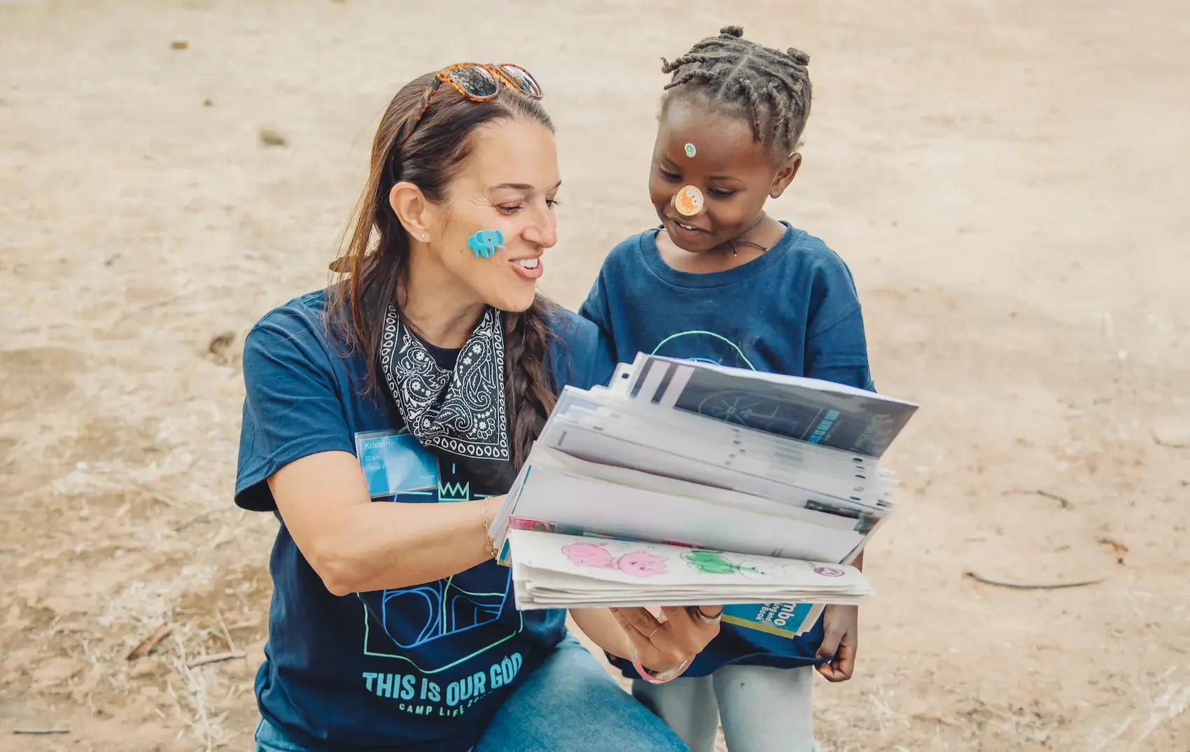 A woman and a young boy looking at a book together outdoors on a dirt ground. The woman has long braided hair and sunglasses on her head, and the boy has short dreadlocks. The woman is smiling while the boy happily looks at the book with him. Stickers decorate both of their faces.