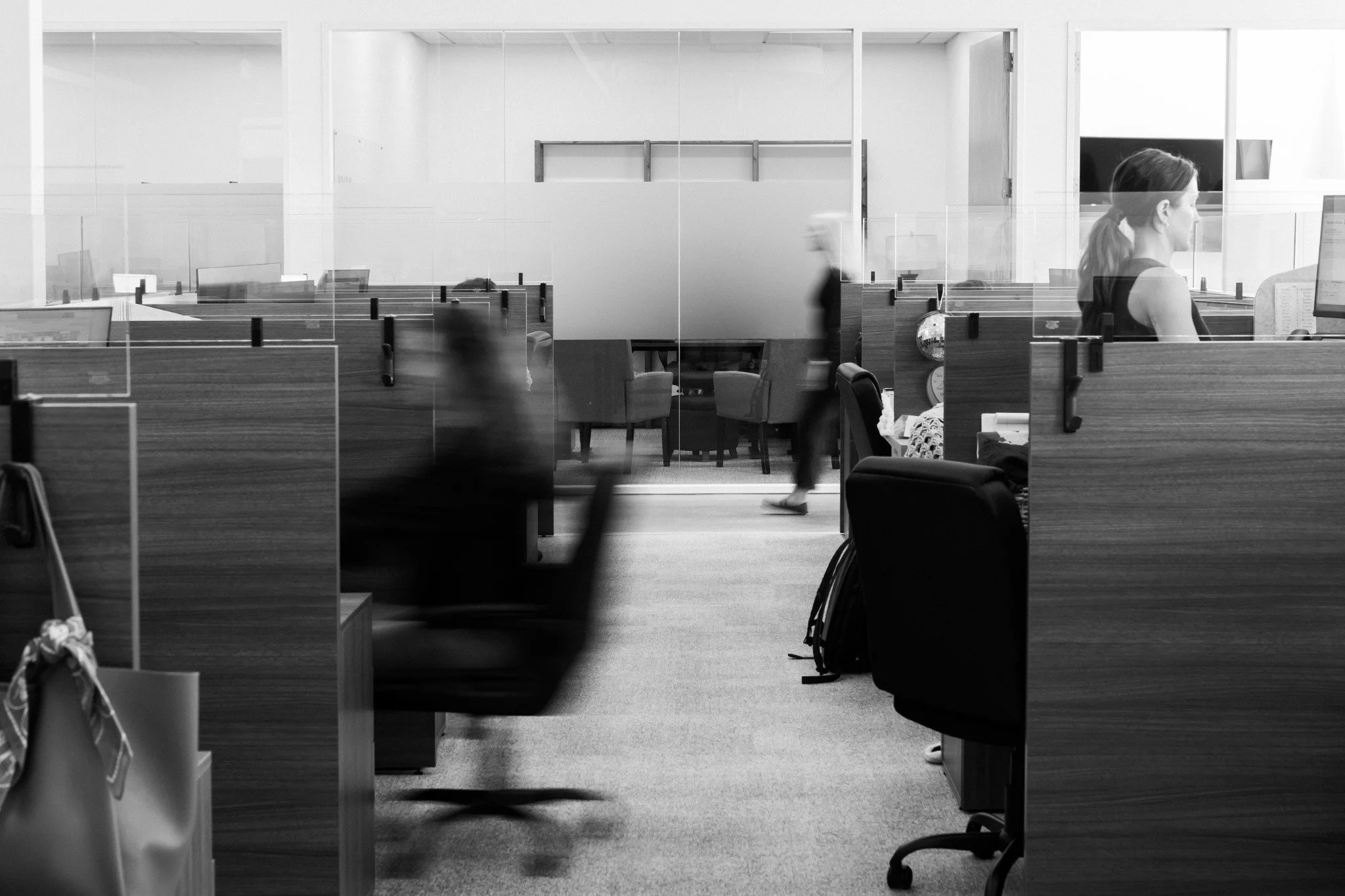 An office cubicle area with a woman working at her desk, seen through a glass wall. The office has multiple cubicles, some chairs, and a closed door in the background.