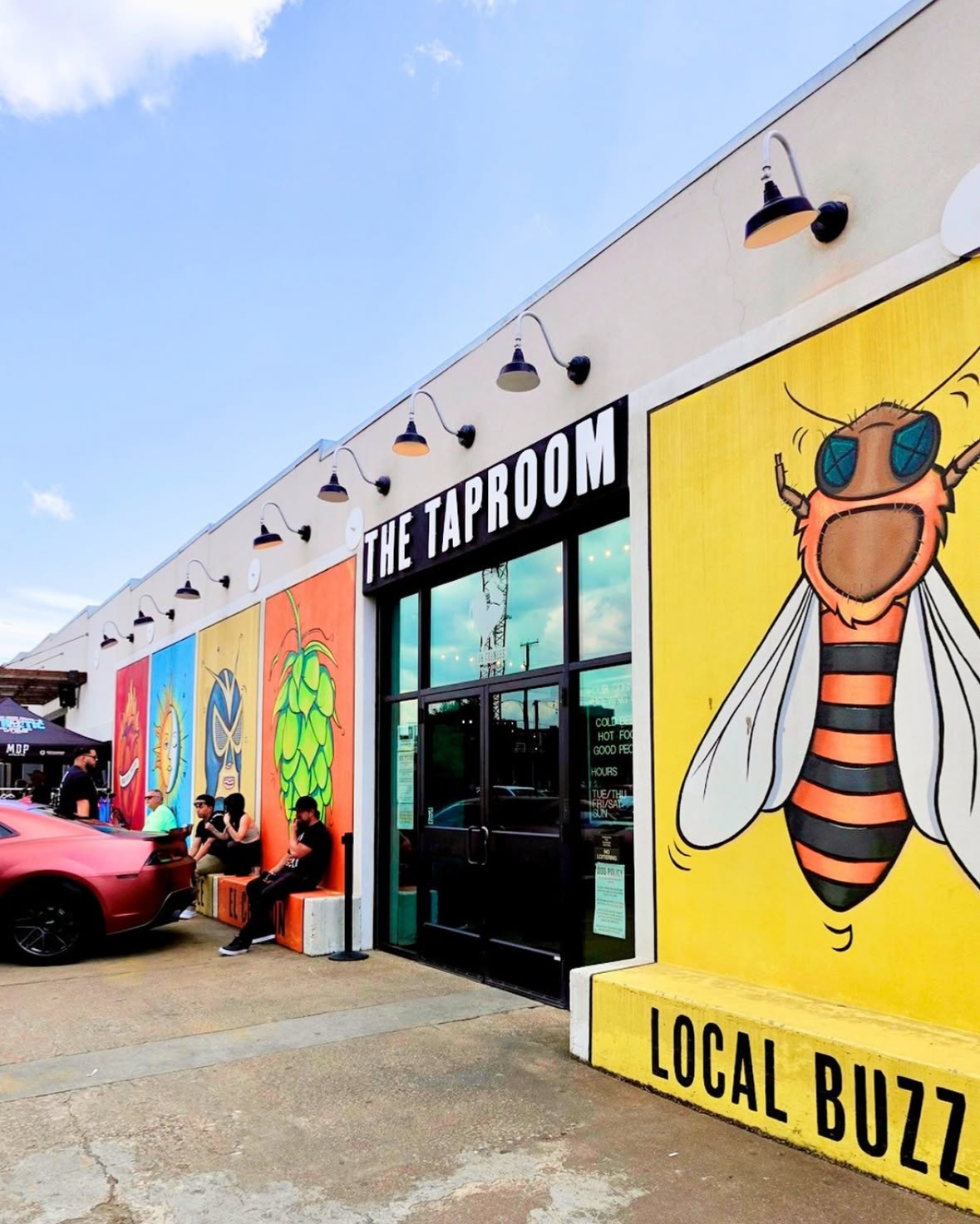 The exterior of a building named 'The Taproom' with colorful murals of a bee and other designs, people sitting outside, and a red car parked nearby. Bright yellow wall with black text reading 'Local Buzz'. Four Corners Brewery, Dallas, Tx