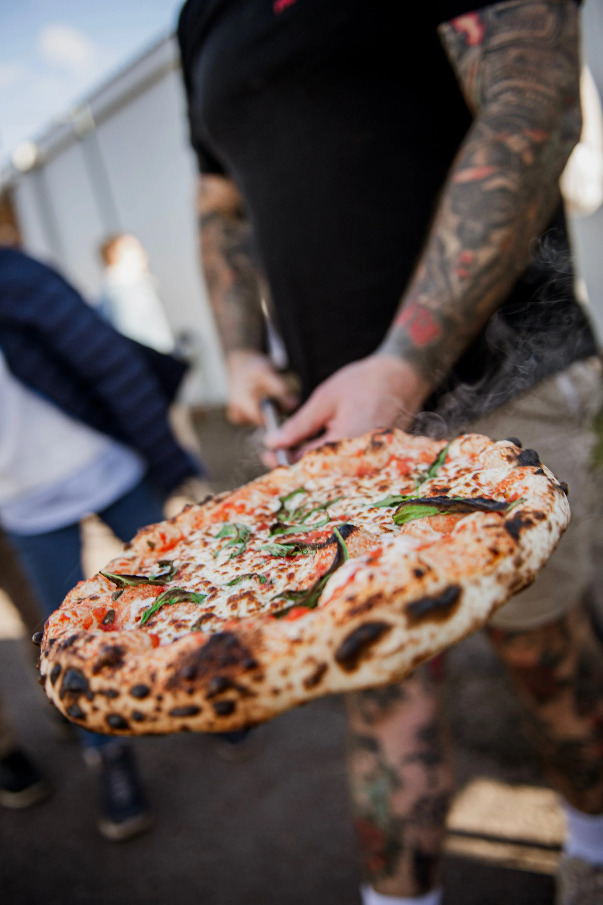 Close-up of a person holding a freshly baked pizza with toppings including basil and cheese, with a thin crust, near a group of people.