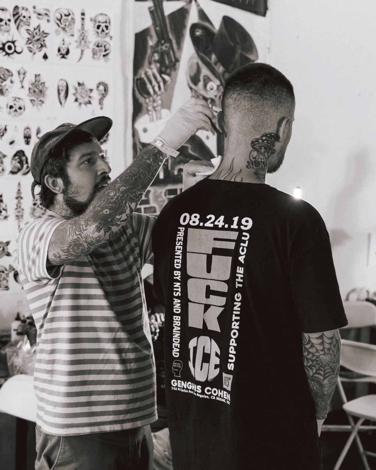 A tattoo artist with tattoos on both arms is applying a tattoo to a young man with a buzz cut, who is wearing a black t-shirt with event information on the back. The background includes tattoo flash art on the wall.