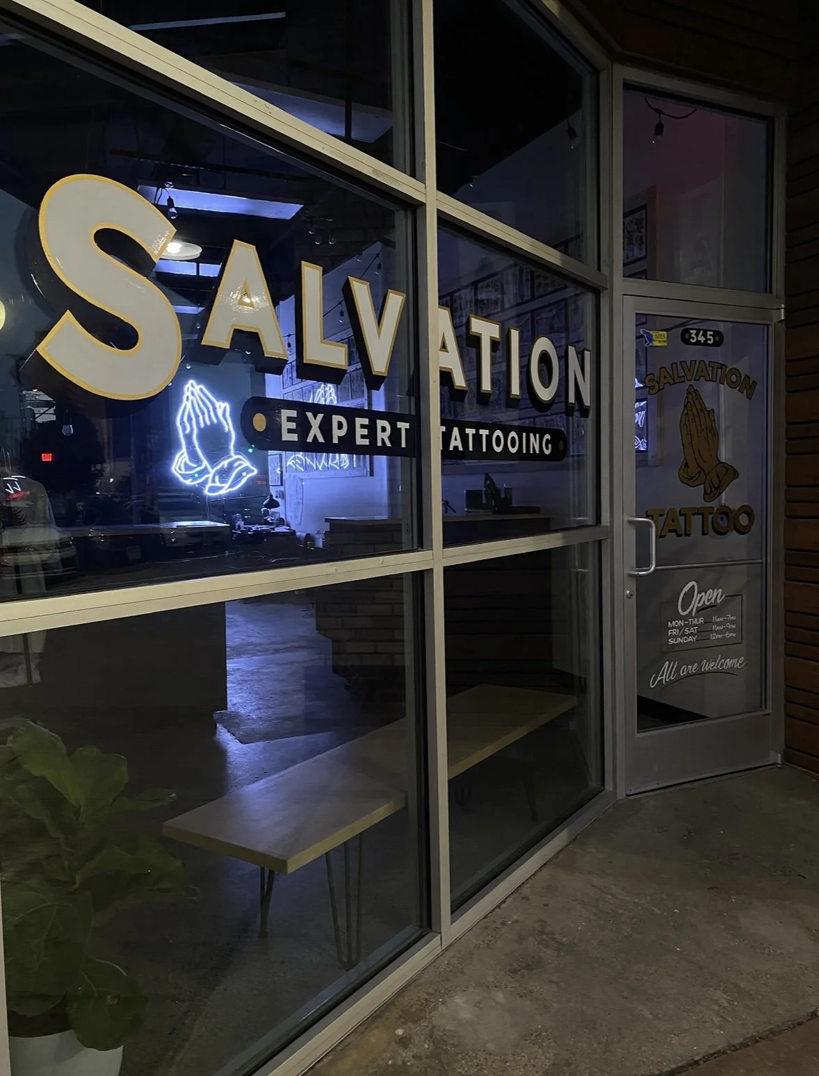 Storefront window displaying the name 'Salvation Expert Tattooing' with a neon praying hands sign, and a door with an 'Open' sign, hours, and the logo of praying hands. Salvation Tattoo, Dallas, Tx