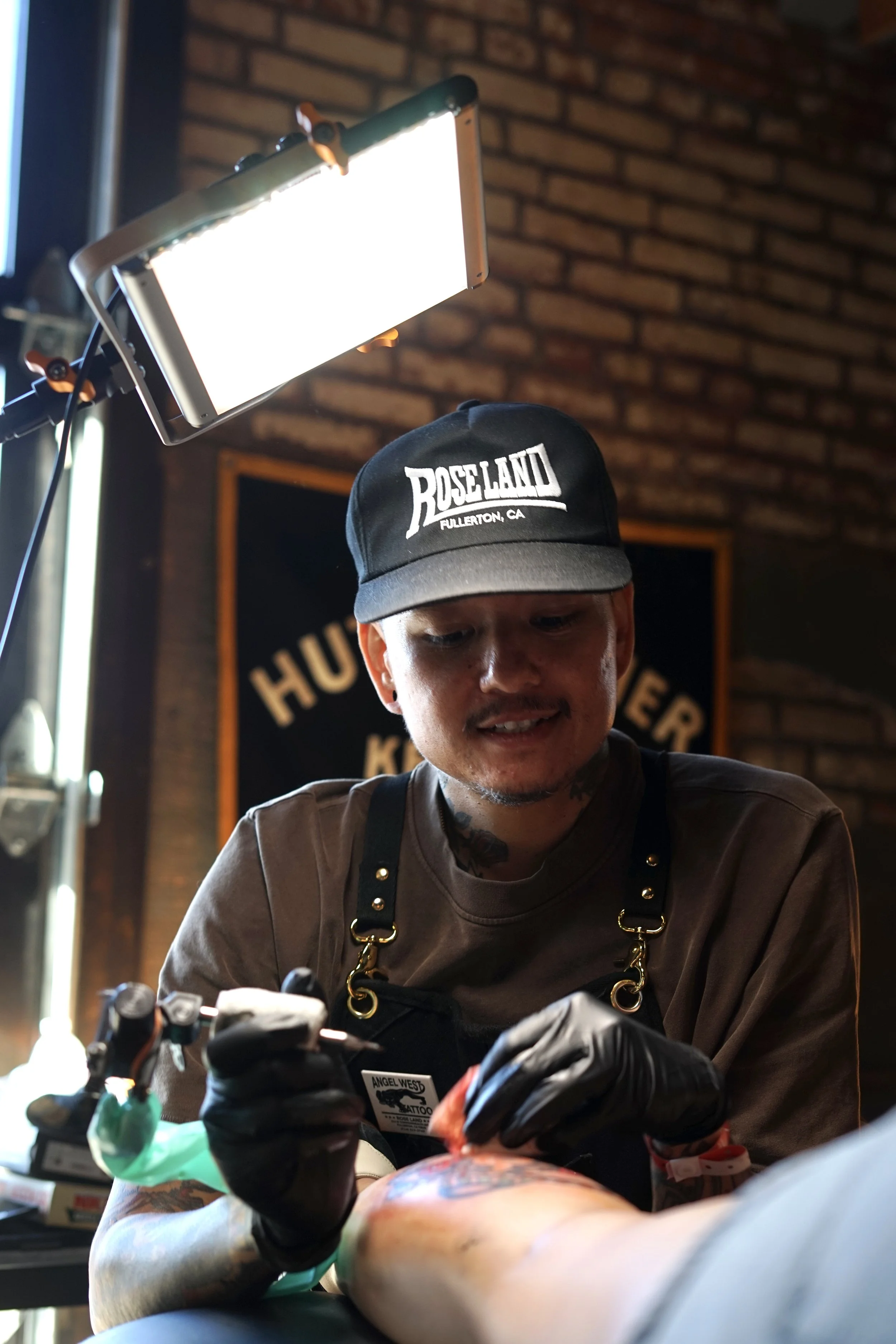 Tattoo artist with black gloves, wearing a black cap with white text, working on a customer's arm in a tattoo studio with brick walls and lighting equipment.