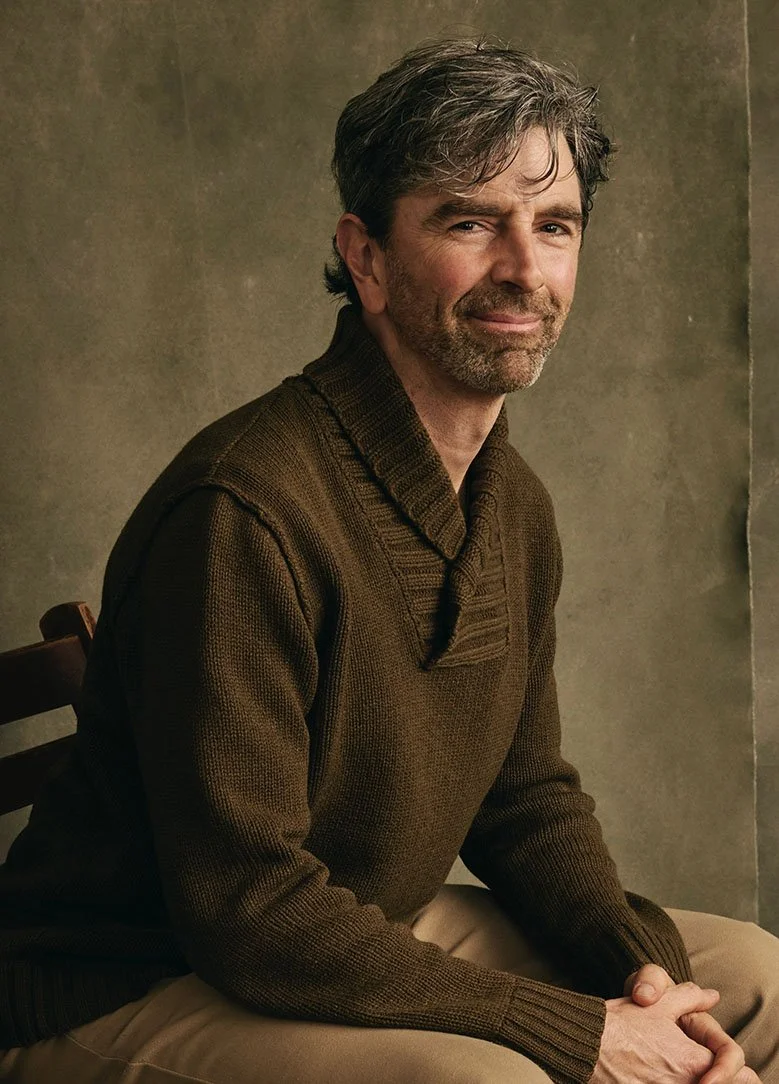 A middle-aged man with grayish hair and beard sitting on a wooden chair against a plain, textured wall, wearing a brown knitted sweater and beige pants, smiling slightly at the camera.