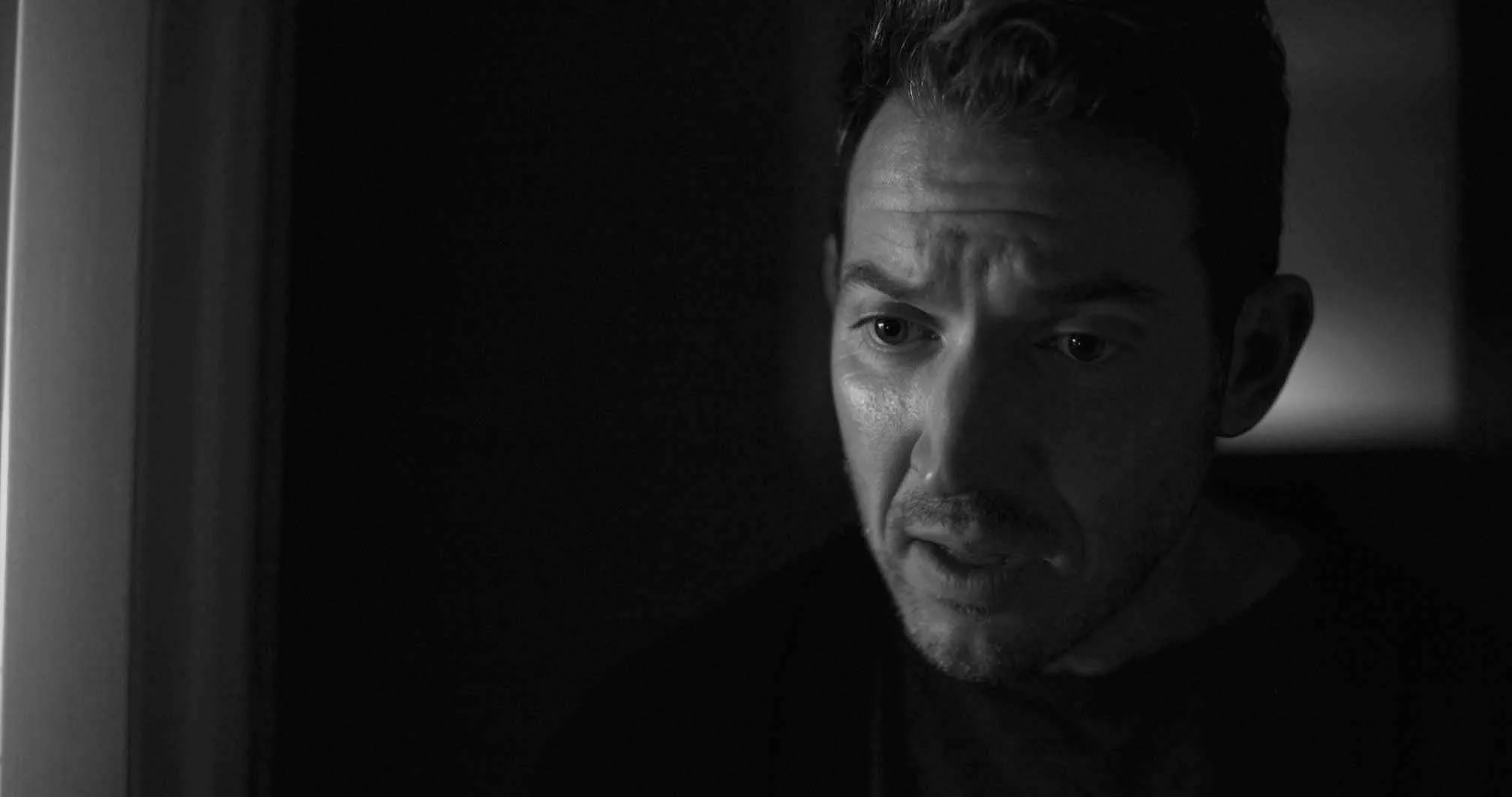 Black and white close-up of a man's face, showing part of his forehead, eye, nose, and mouth, with a serious expression, looking slightly downward.