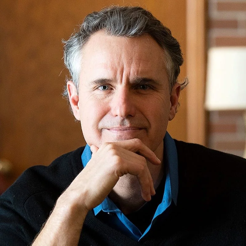 A middle-aged man with gray hair, wearing a dark sweater over a blue collared shirt, sitting indoors with a thoughtful expression, resting his chin on his hand.