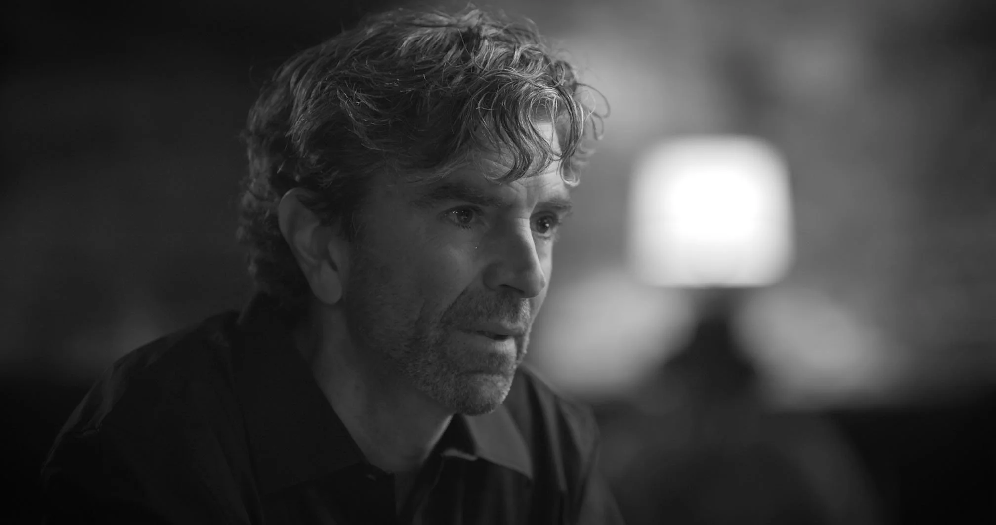 A black and white close-up of a middle-aged man with wavy hair, tear rolling down his cheek, looking thoughtful or distressed, with a blurred background.
