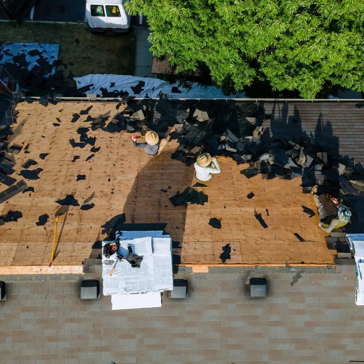 Aerial view of roof replacement process with workers removing damaged shingles and preparing residential roof decking for new installation.
