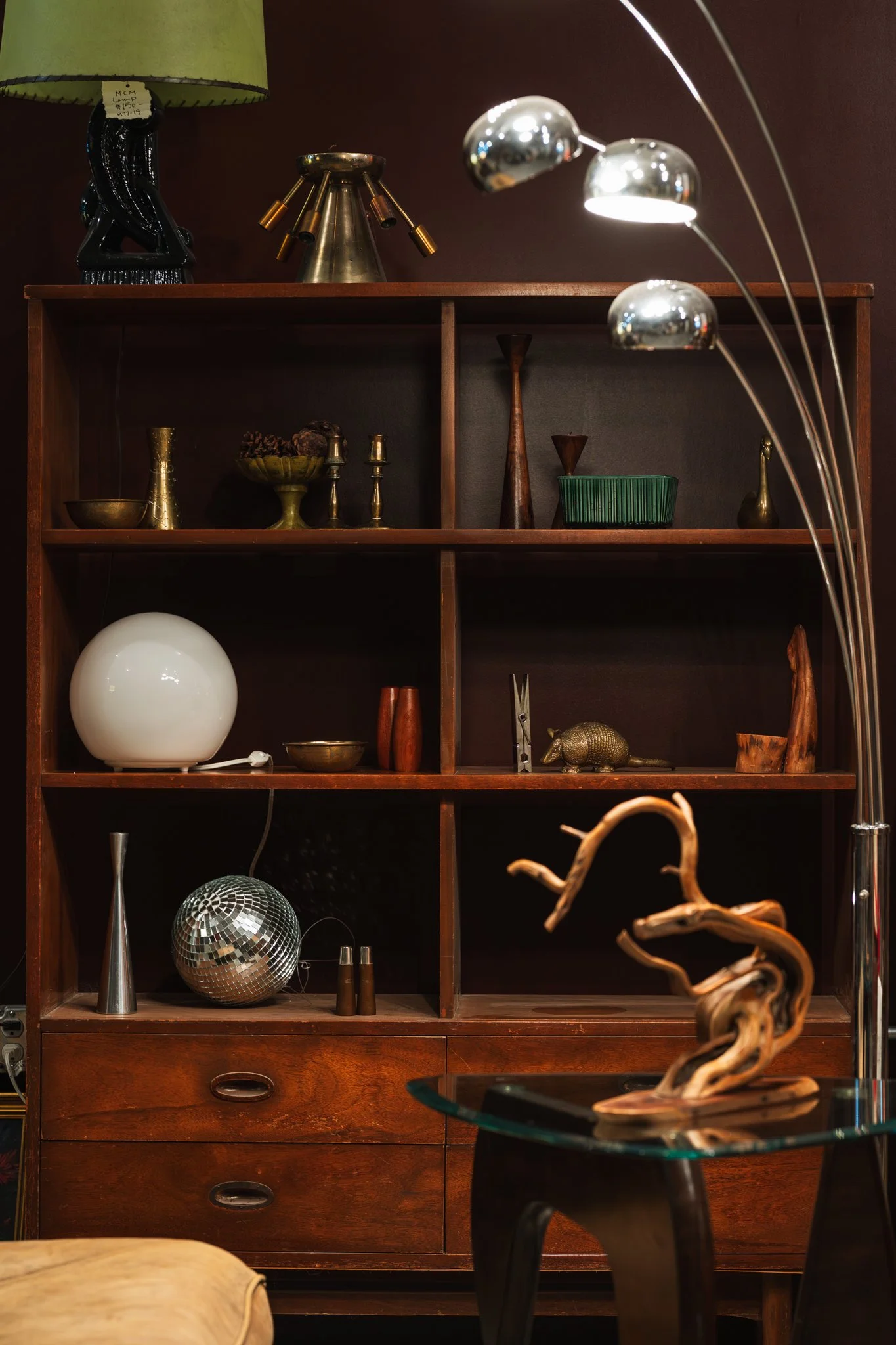 A wooden bookshelf with various decorative items, including a white spherical lamp, a metallic disco ball, and a wooden sculpture resembling a tree branch, in a room with dark walls.