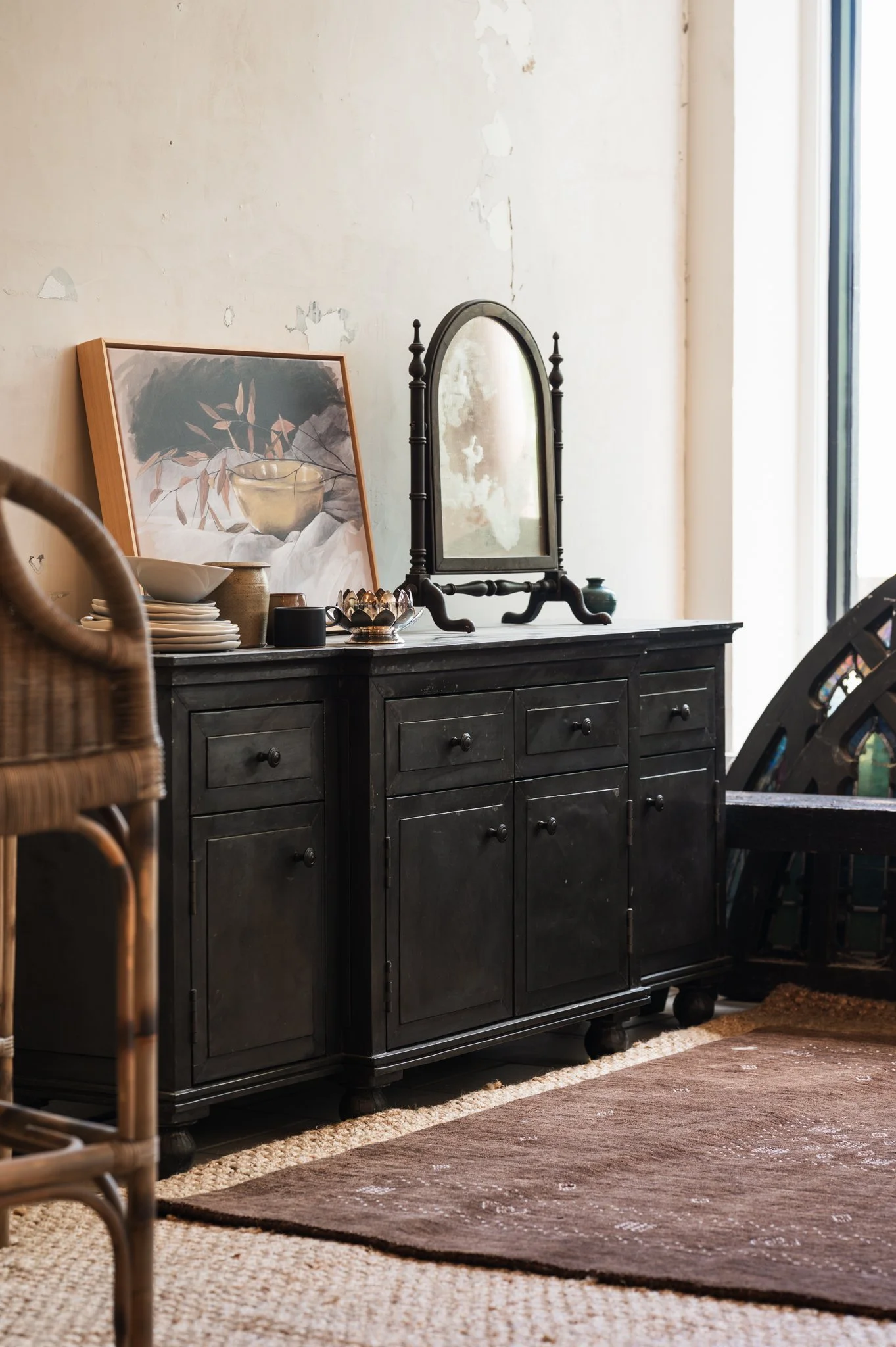 A black wooden sideboard with a mirror, painting, and tableware on top, set against a wall with peeling paint, next to a window with sunlight.