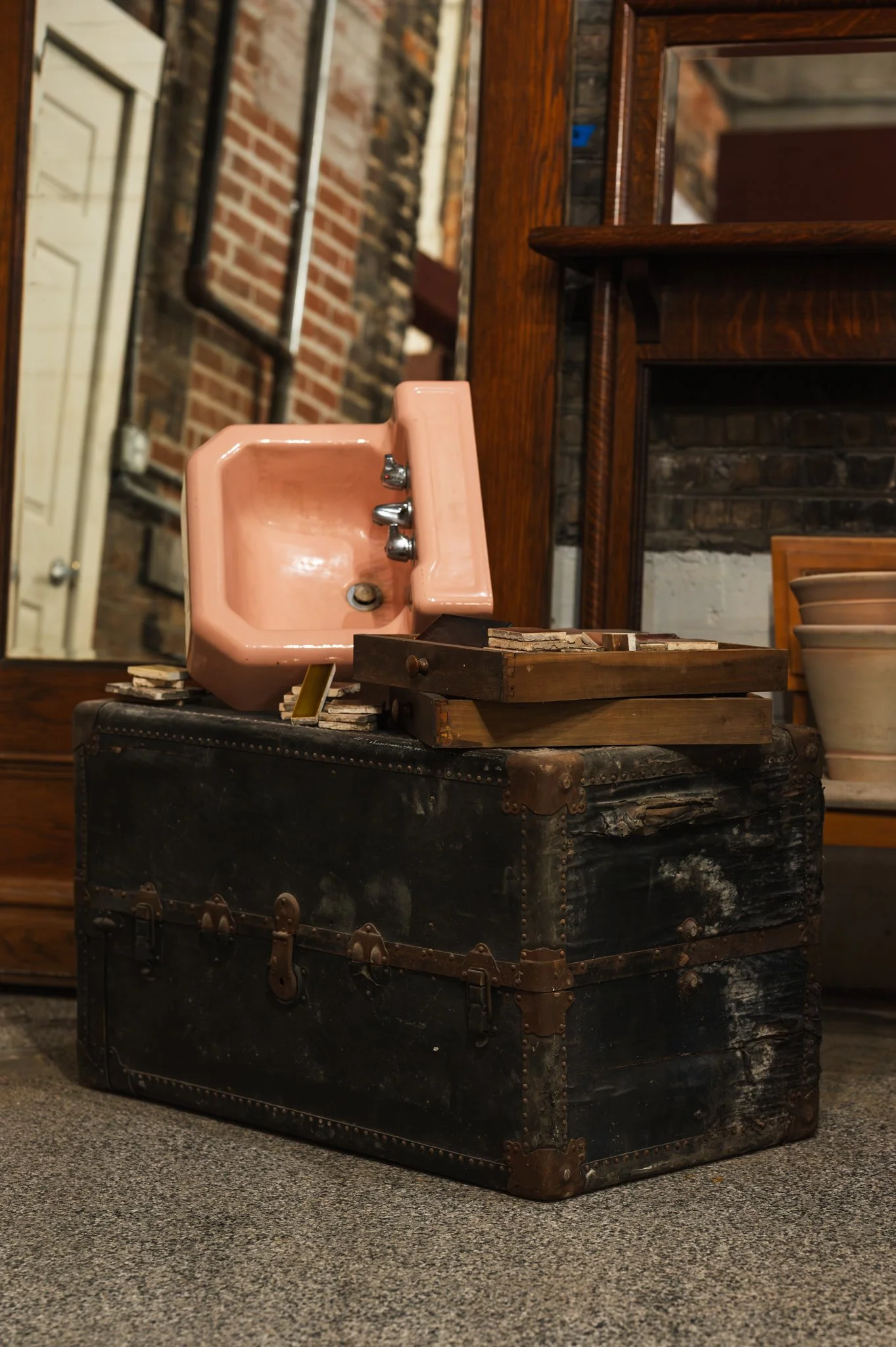 A vintage black trunk with iron accents and a worn surface, holding a stack of beige pots and a wooden crate with small tiles, with a pink ceramic sink and a wooden cabinet against a brick wall in the background.