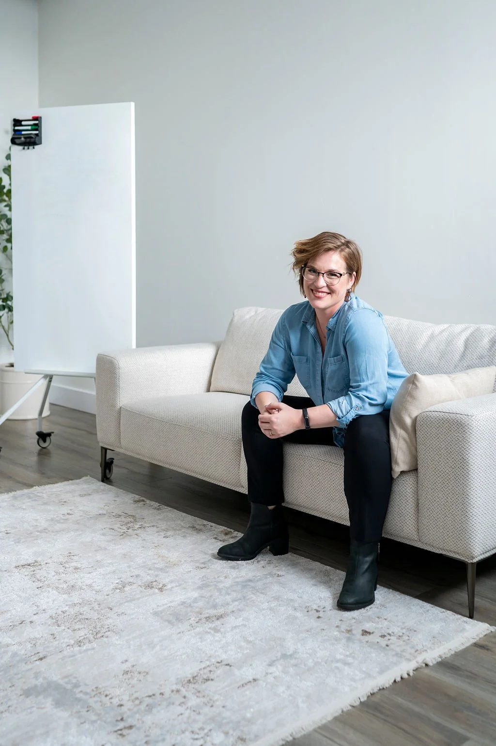A woman with short hair, glasses, wearing a denim shirt, black pants, and black ankle boots, sitting on a white sofa in a modern, minimalistic room with grey walls and a white rug.
