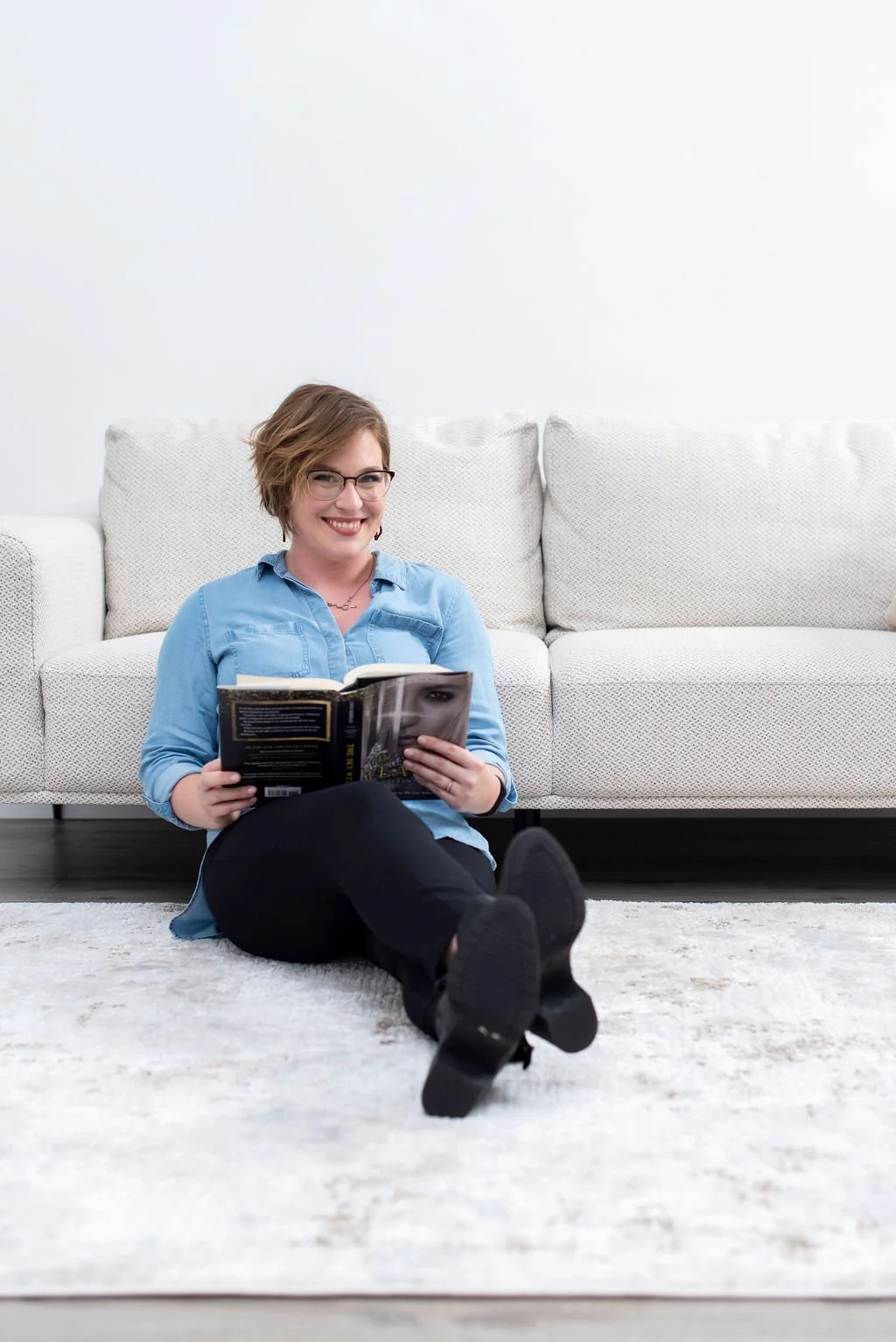A woman with short brown hair, glasses, and a blue shirt sitting on a white rug, smiling and reading a book in a bright, minimalistic living room with a white couch and pillows.