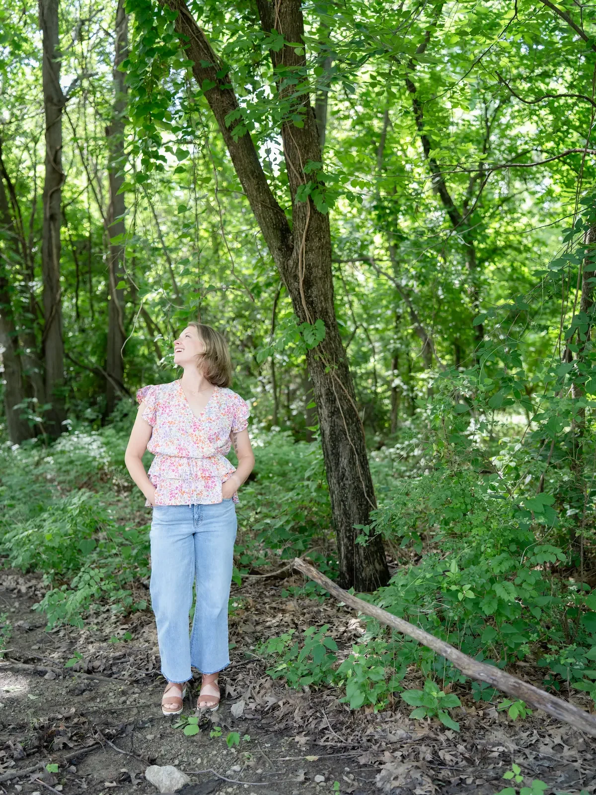 A female therapist stands on a wooded trail, smiling and looking upward. She has short brown hair and is wearing a pink floral top, light blue jeans, and sandals. The background is filled with green foliage and tree trunks.