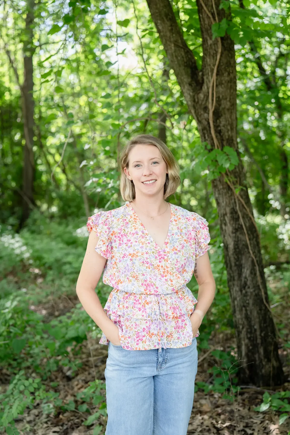 A female therapist with shoulder-length blonde hair smiling outdoors in a wooded area, wearing a floral short-sleeve top and light blue jeans.