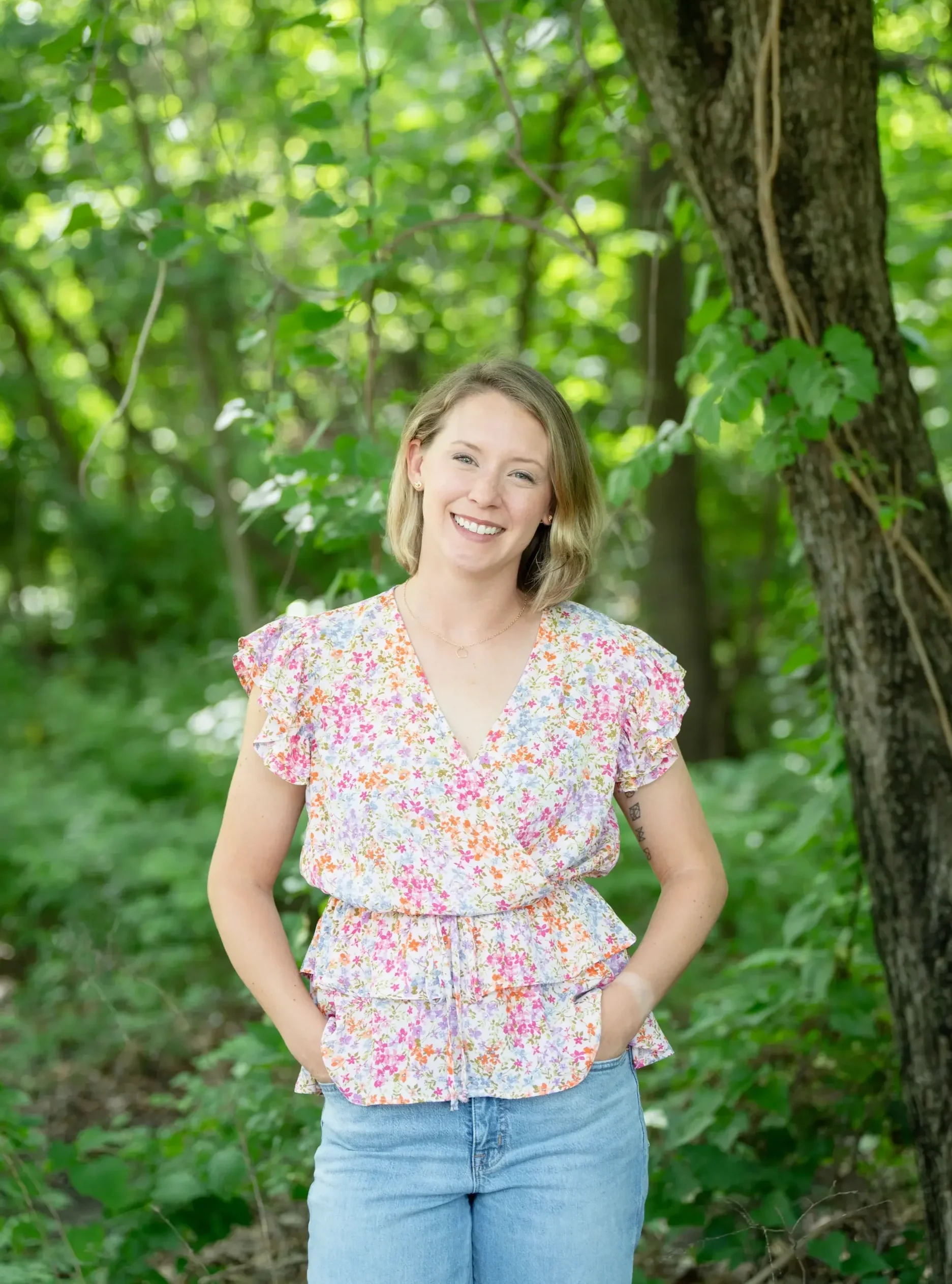 A smiling female therapist with blonde hair standing outdoors in a green, wooded area. She is wearing a floral top and blue jeans.