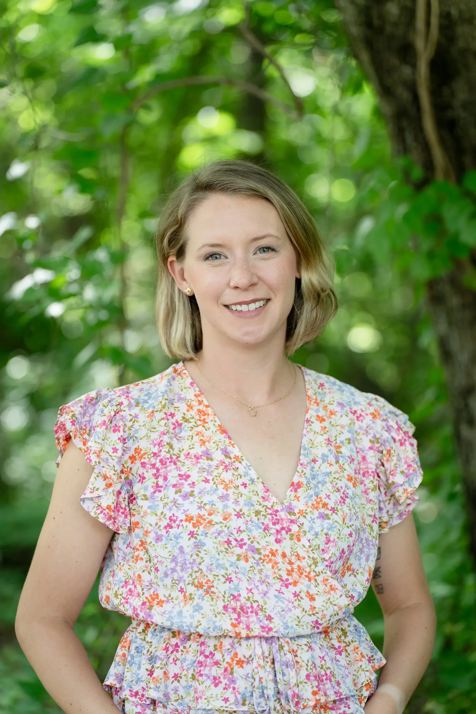A female therapist with short blonde hair, wearing a floral dress, smiling outdoors in a green, leafy setting.