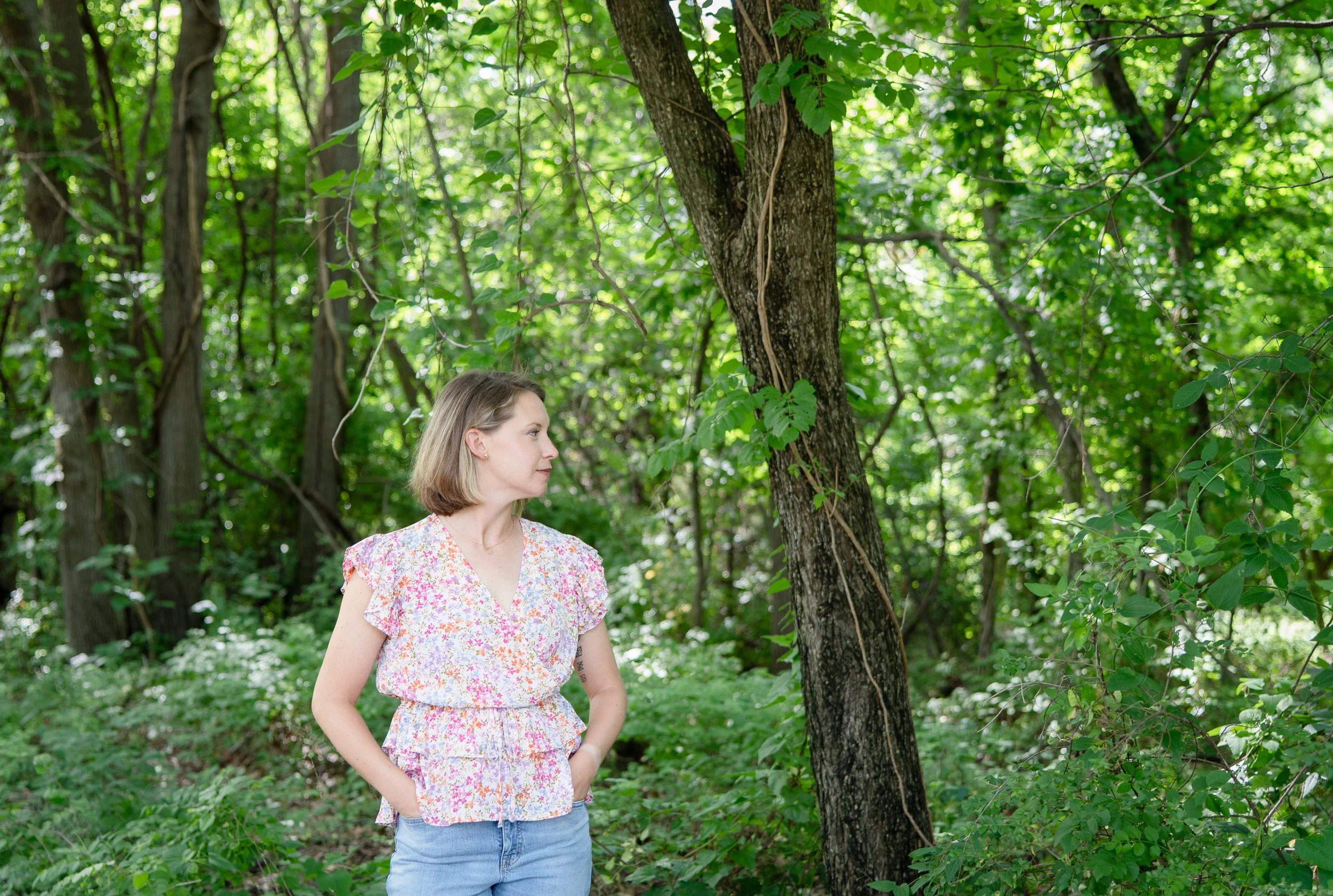 A  female therapist with blonde hair wearing a floral dress standing in a wooded area with green foliage and trees.