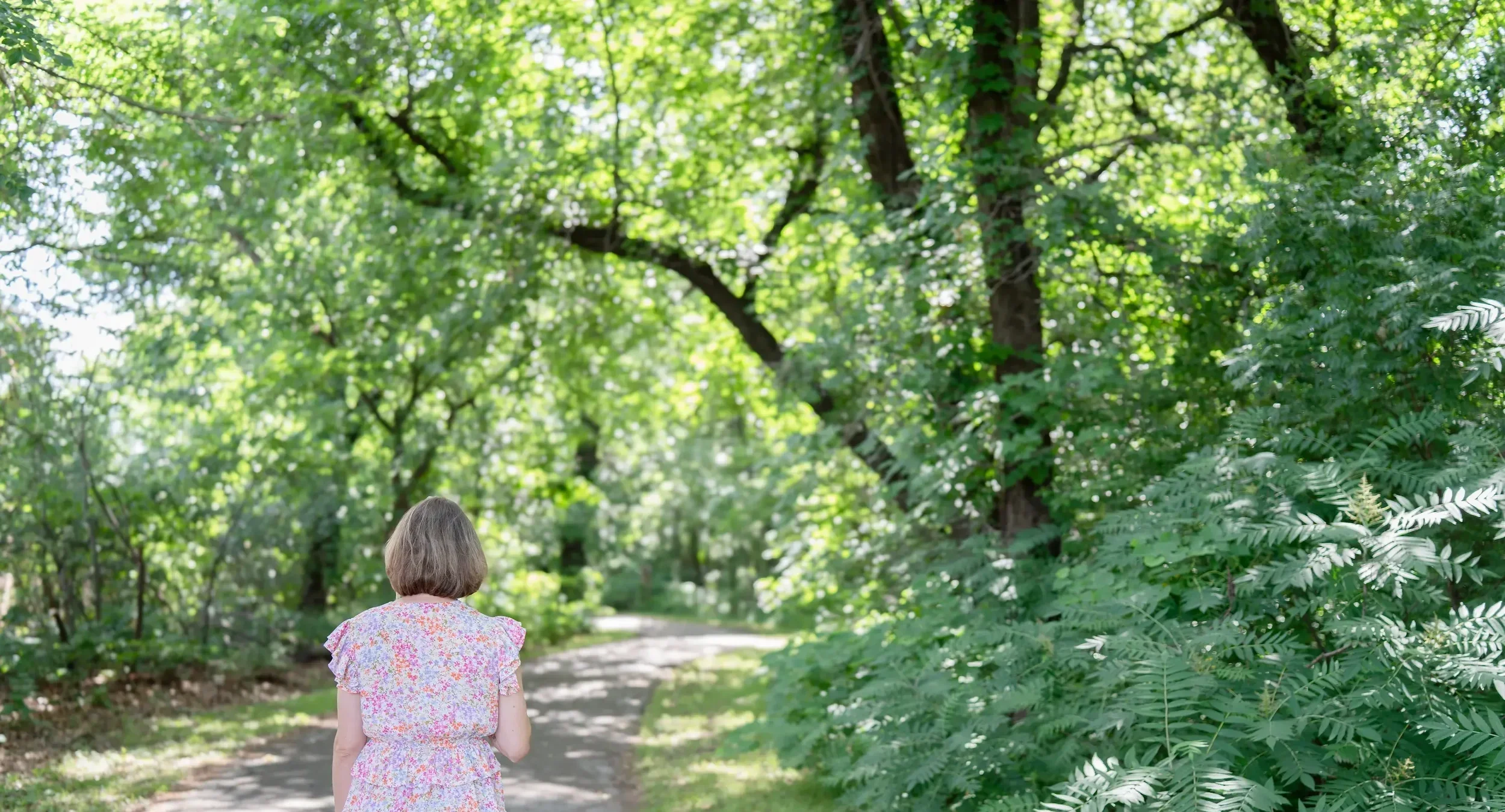 A  female therapist with shoulder-length hair, wearing a pink floral dress, walking on a dirt path in a lush green forest canopy with sunlight filtering through the leaves.