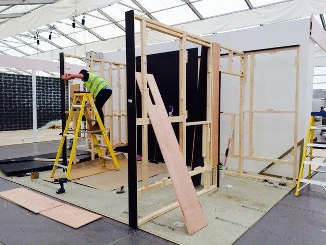 Person in a safety vest working on construction site, standing on a ladder building a structure with wooden frames inside a tent.