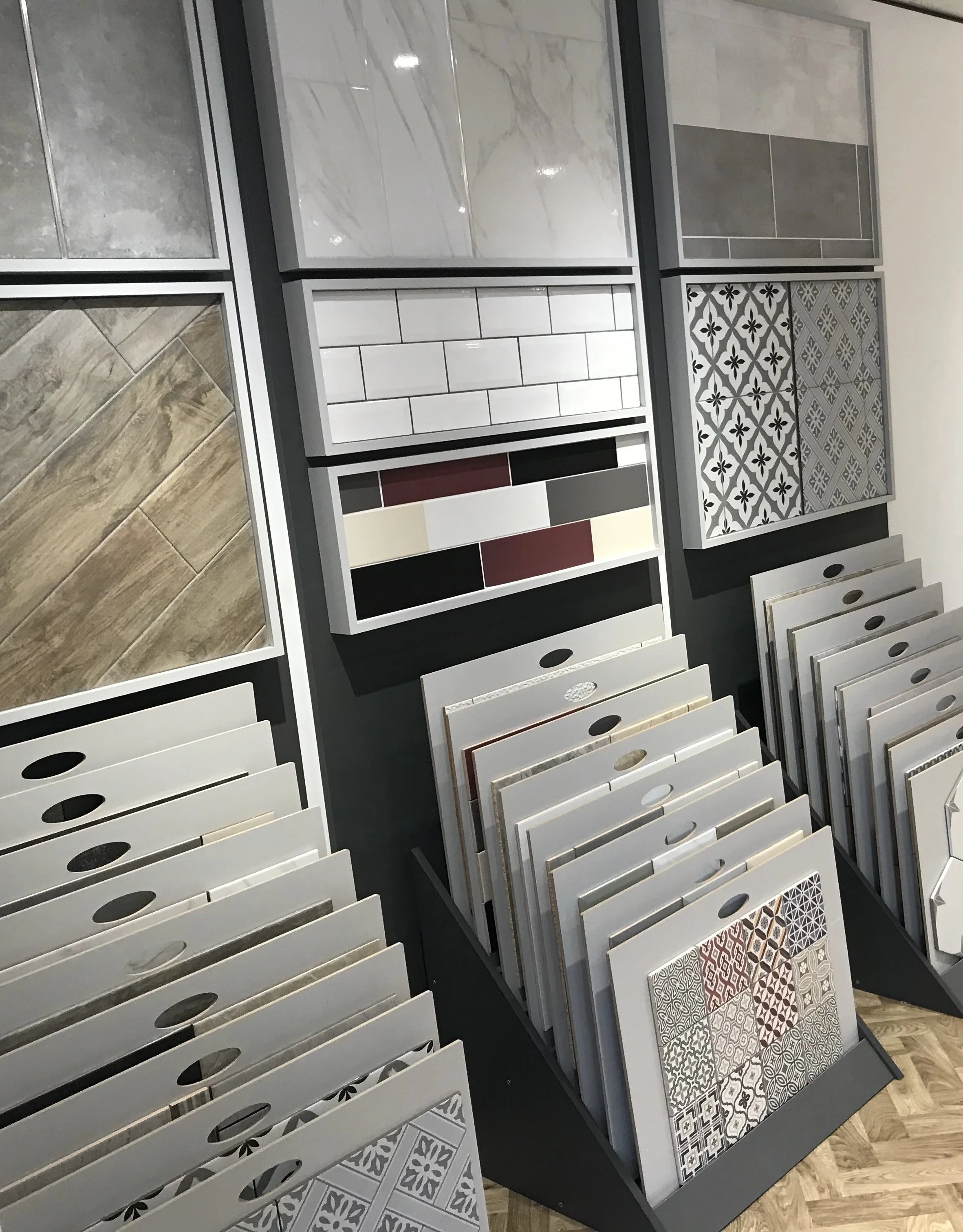 Display of various tile samples in a showroom, featuring different designs and colors, including wood-like, marble, subway, and patterned styles.