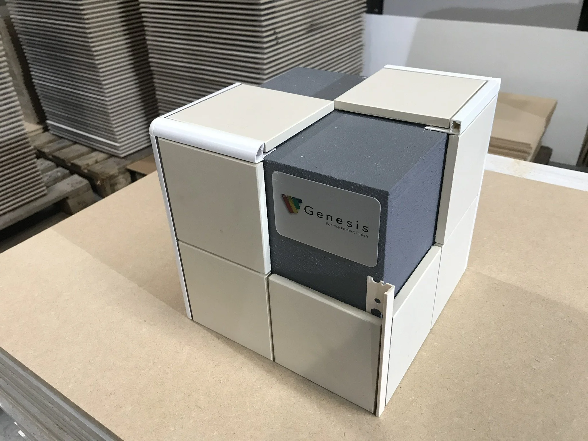 A modular display cube with a "Genesis" logo, showcasing tiled construction materials on a plywood surface.