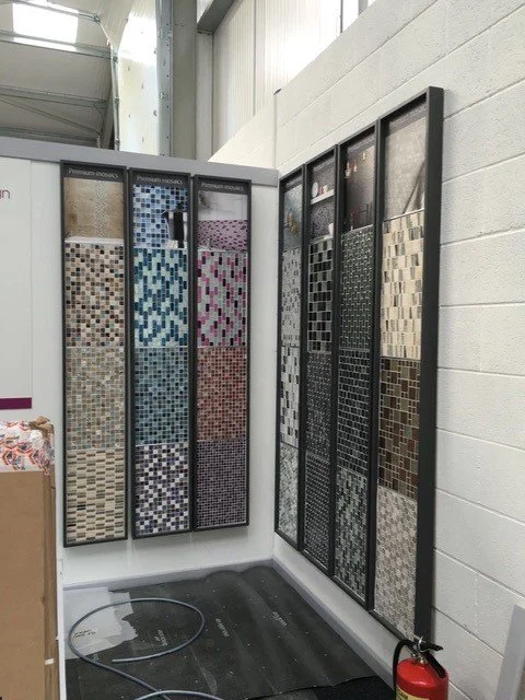 Display of various mosaic tile samples in a showroom with different patterns and colors, against a white wall.
