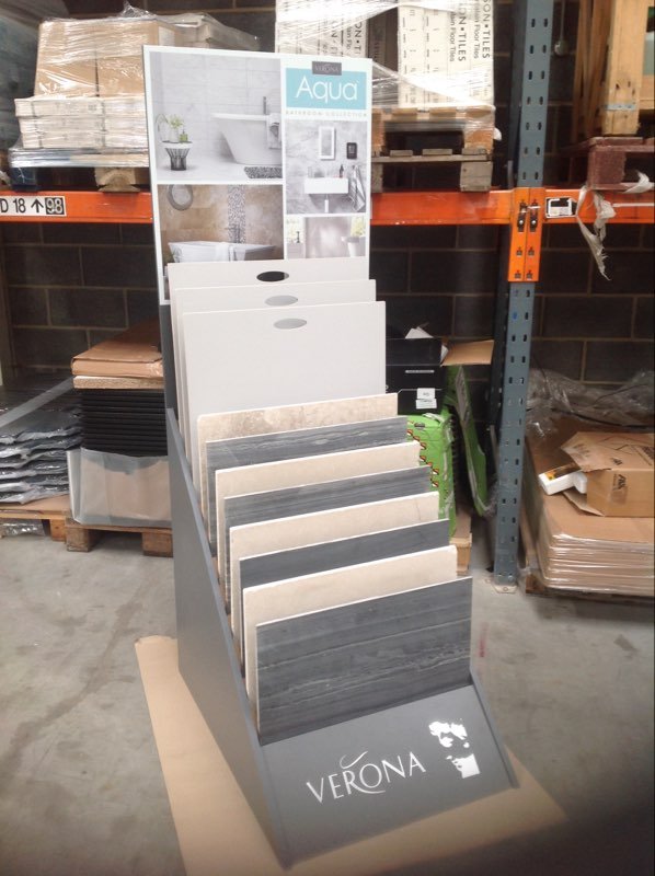 Display stand with various tile samples and promotional sign in a warehouse setting.