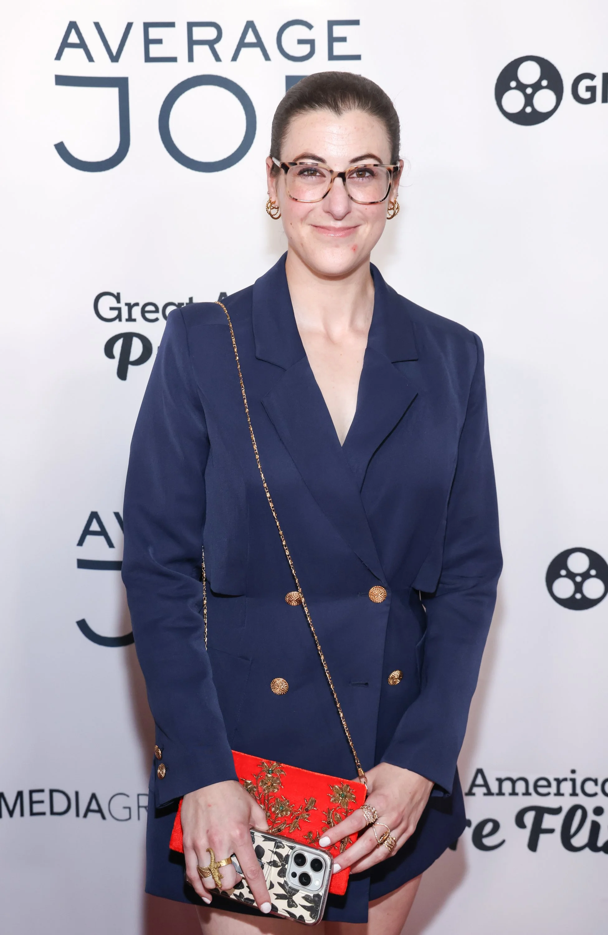 Woman with dark hair pulled back, wearing glasses and gold hoop earrings, dressed in a navy blazer-style dress with gold buttons, holding a red embroidered clutch and a phone, in front of a white backdrop with black text and logo that says 'Average Joe' and 'Great American'.