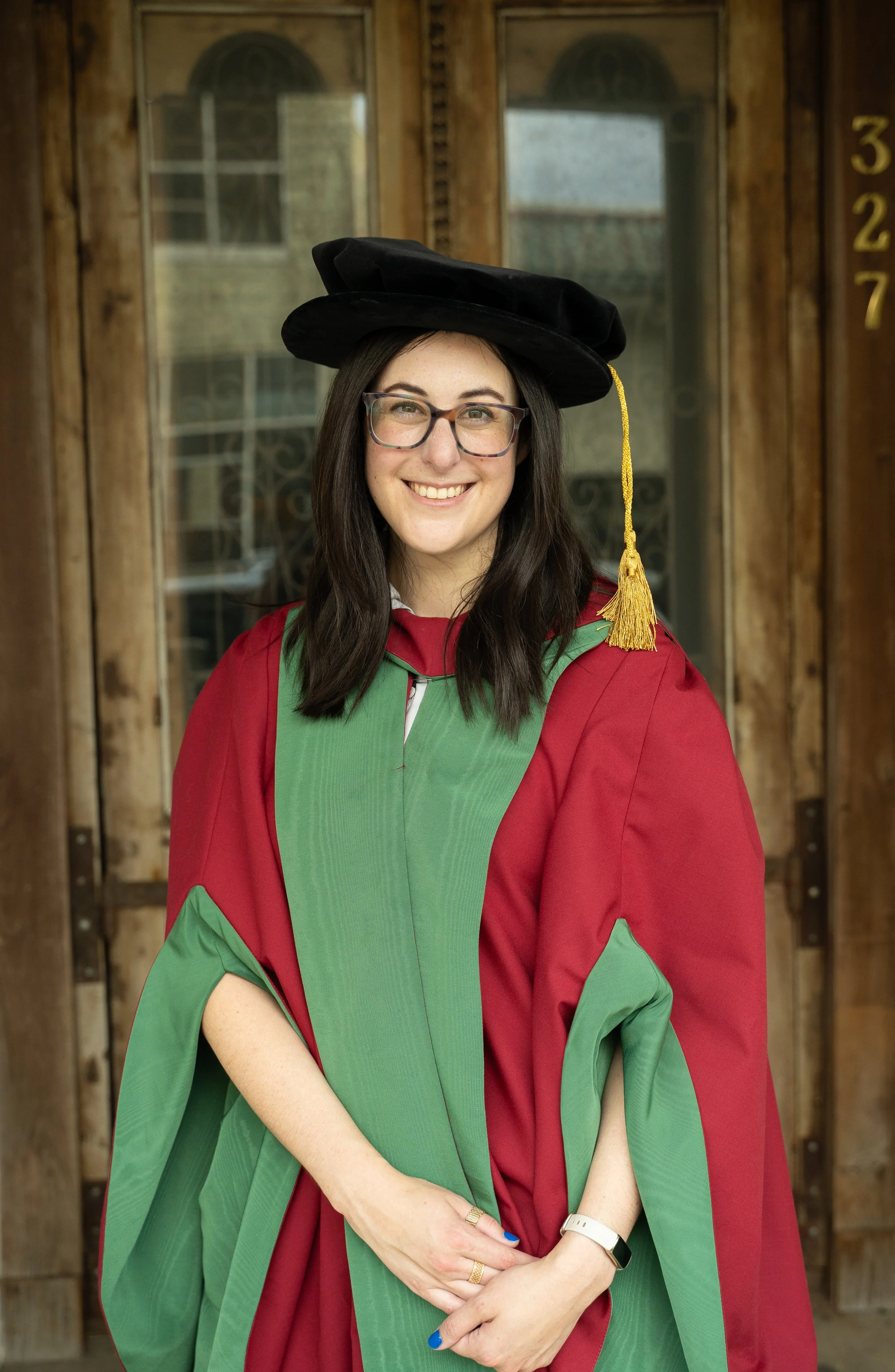 A young woman in graduation regalia, wearing a doctoral cap with a tassel, smiling in front of a wooden door.