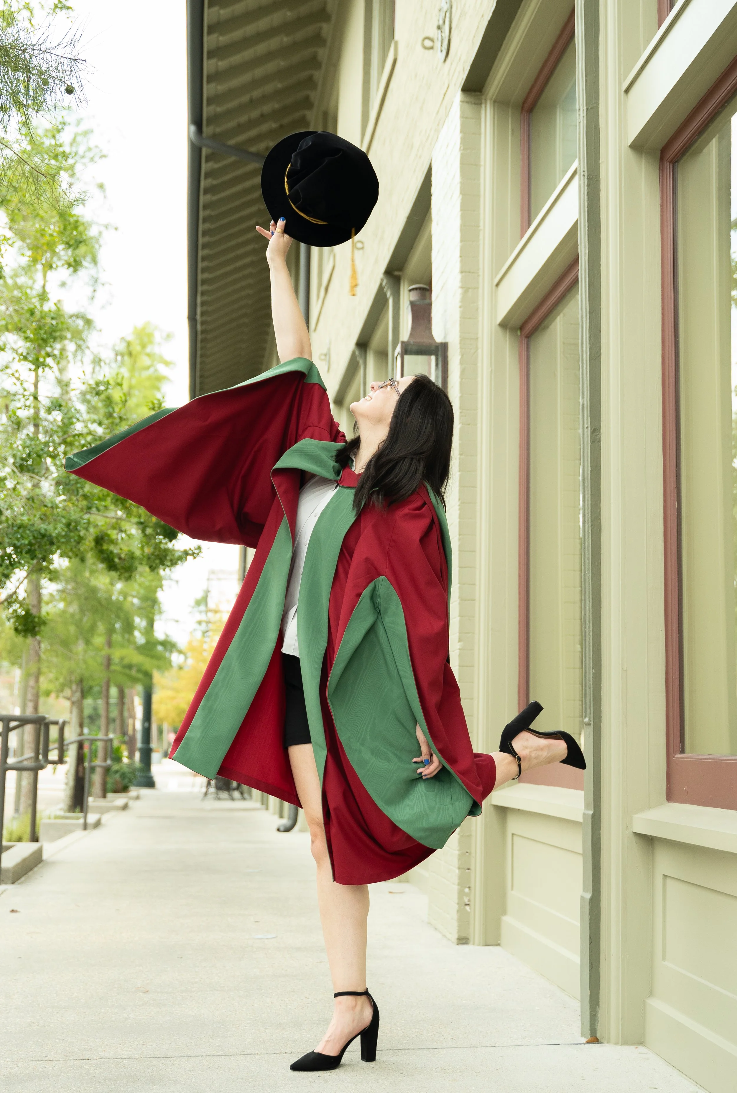 Woman in a graduation gown and heels, standing on one leg with the other leg bent at the knee, holding a hat in the air outside a building with green walls and large windows, green trees and a sidewalk in the background.