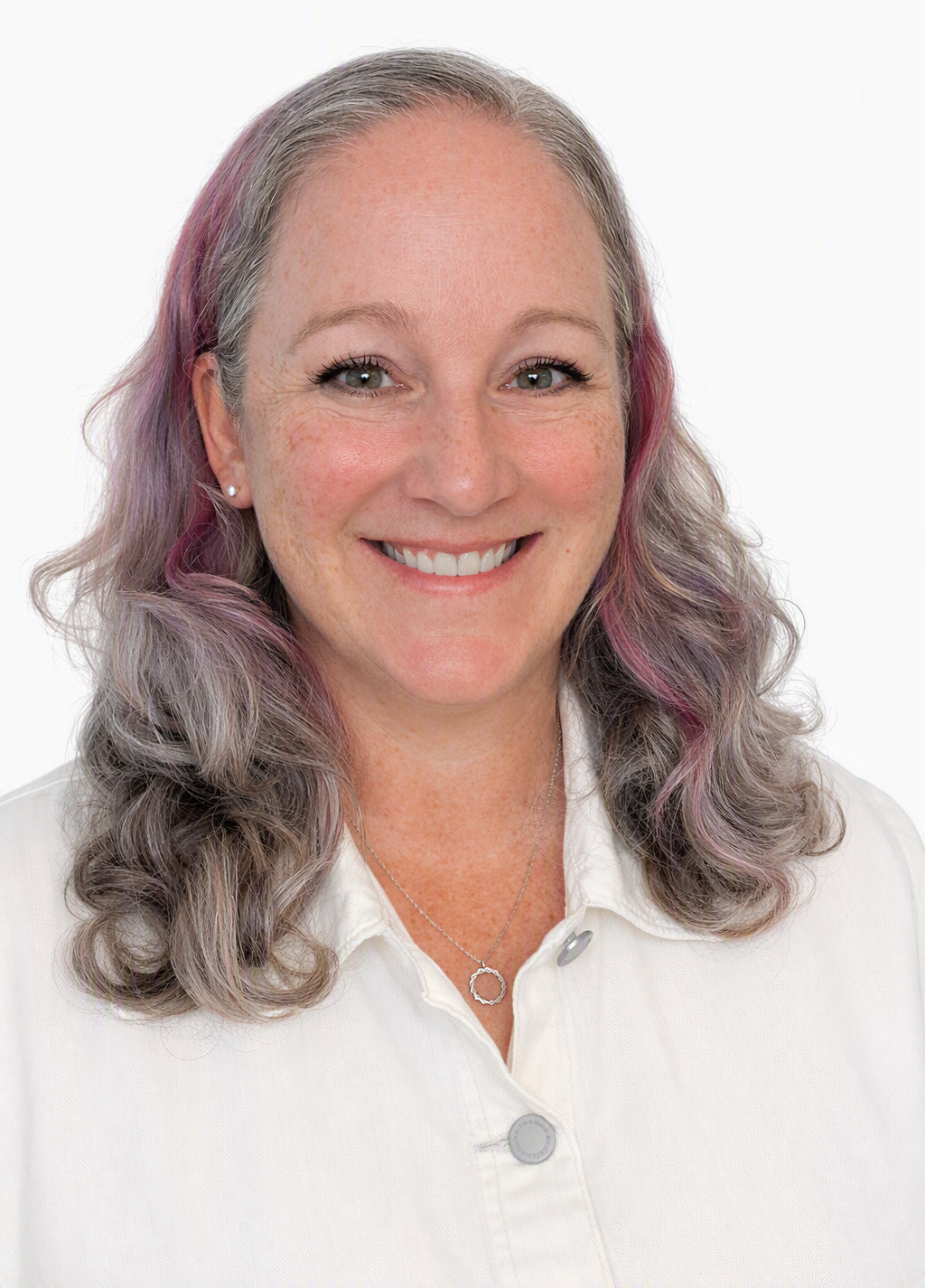 A nurse practitioner with long, wavy, purple and gray hair, smiling, wearing a white shirt, silver necklace, and earrings, against a plain white background.