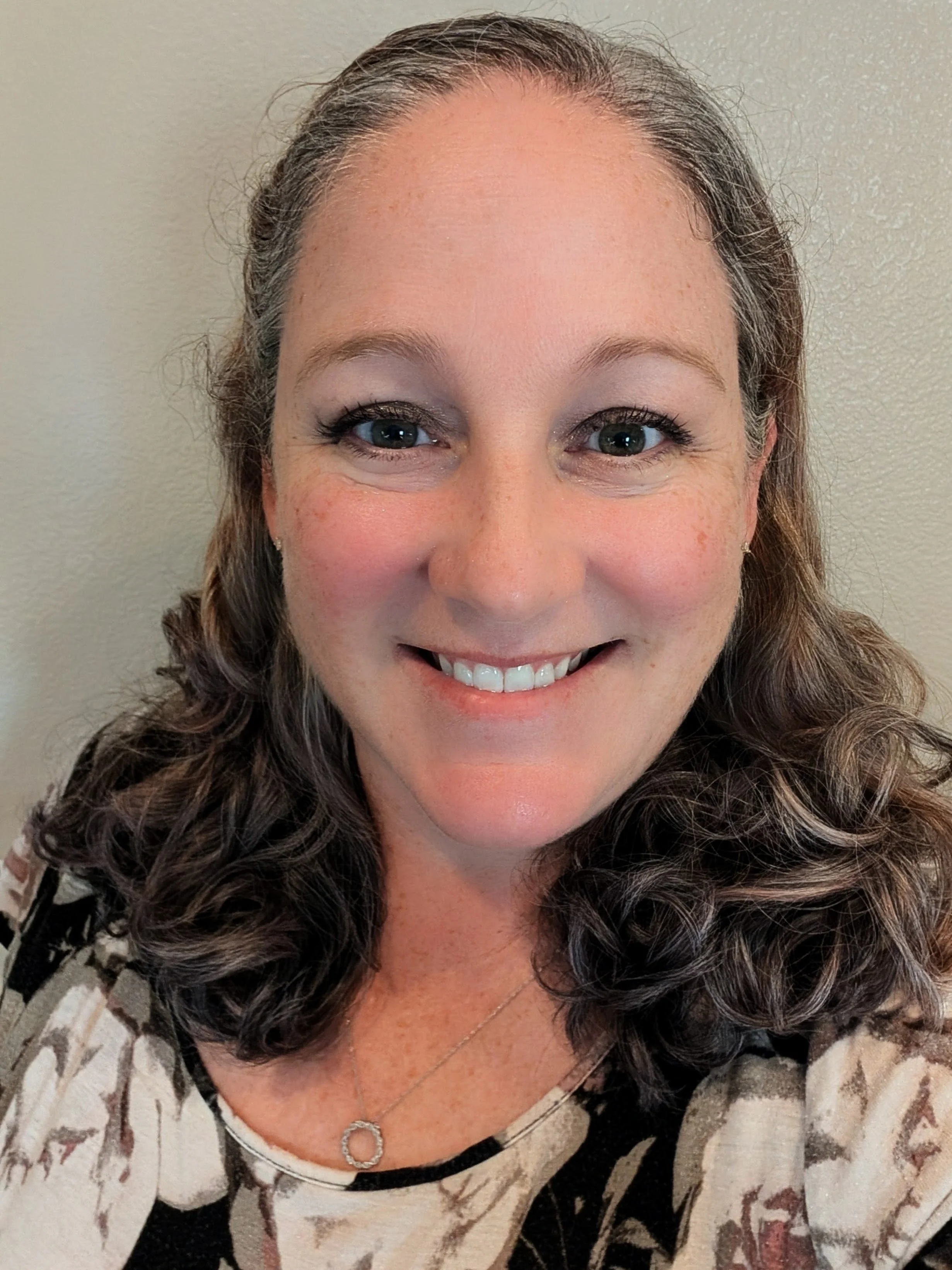 Close-up photo of a smiling woman with curly brown hair, blue eyes, and wearing a patterned top and a silver pendant necklace.