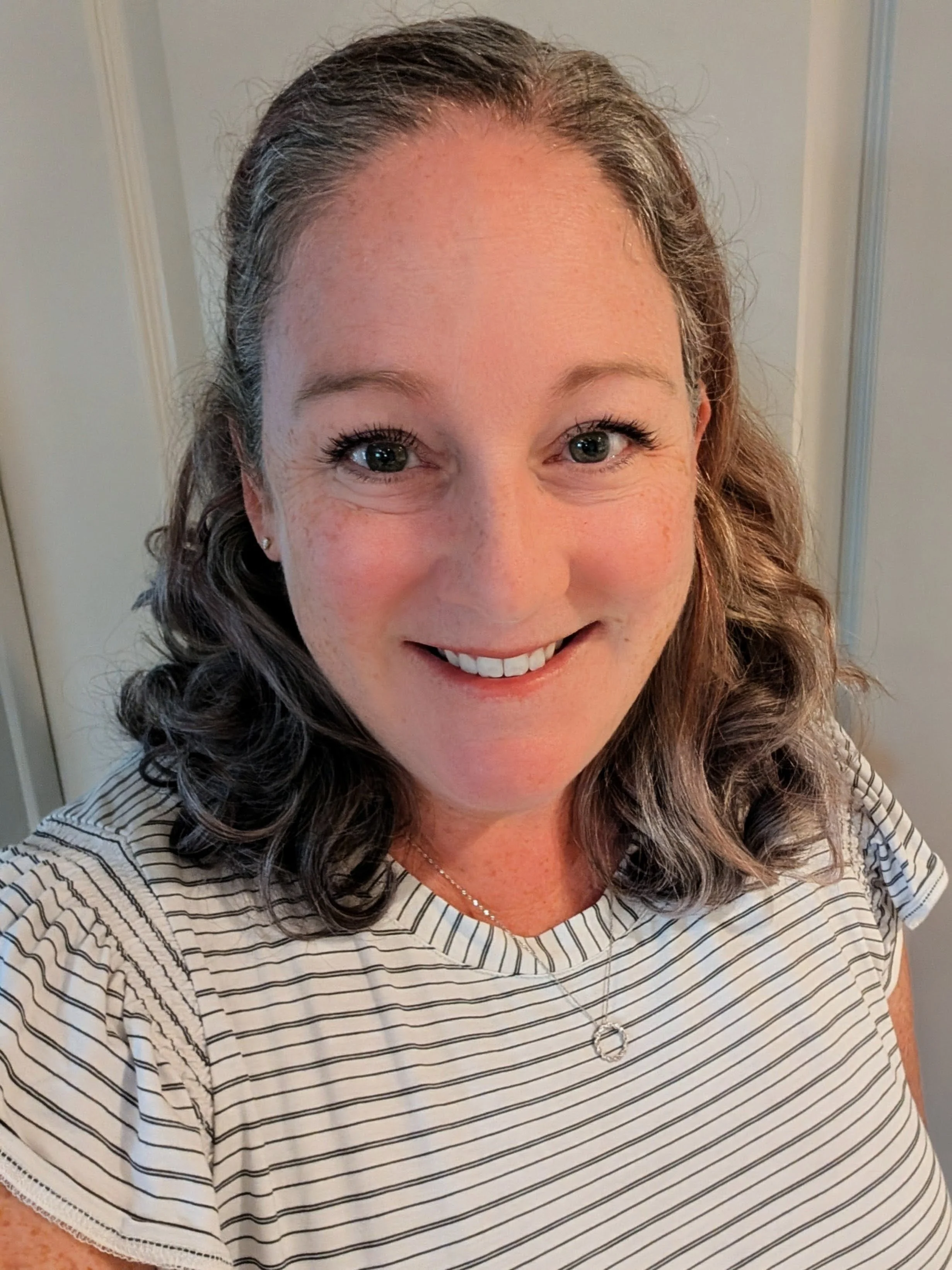 Close-up of a smiling woman with curly brown hair and green eyes, wearing a striped shirt and a silver necklace, standing indoors.