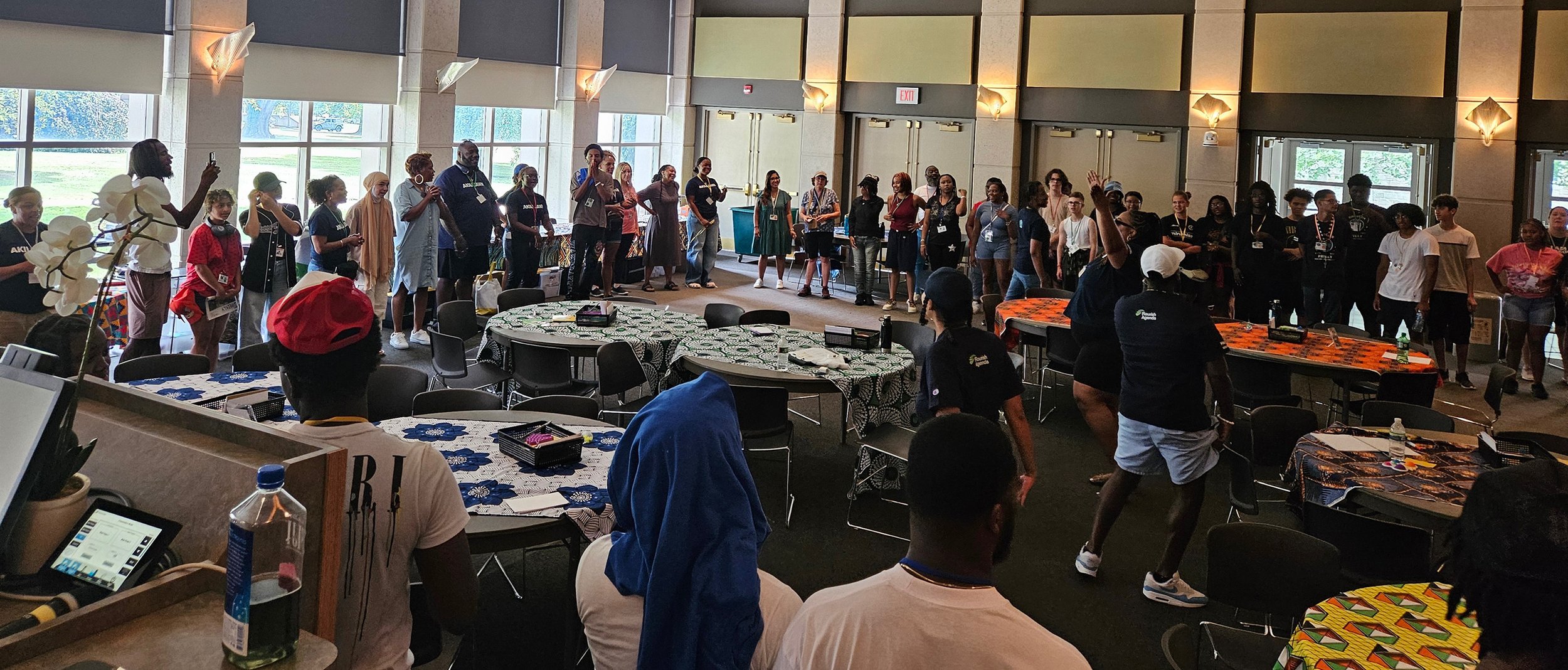 Group of diverse people standing in a large conference room, some with hands raised, participating in a group activity or discussion.